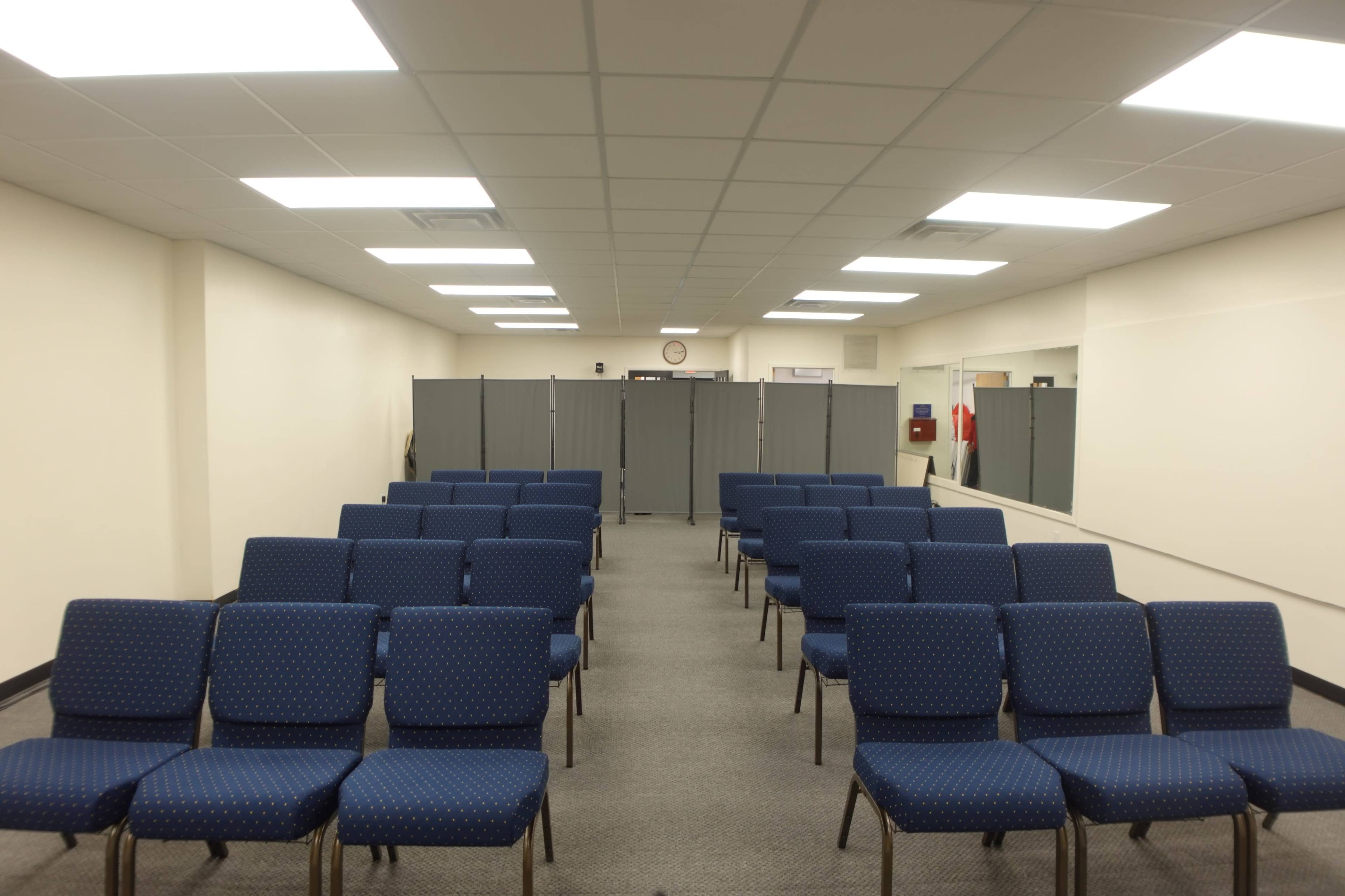 A room with rows of blue chairs arranged in a seating layout, facing a partitioned area at the back.