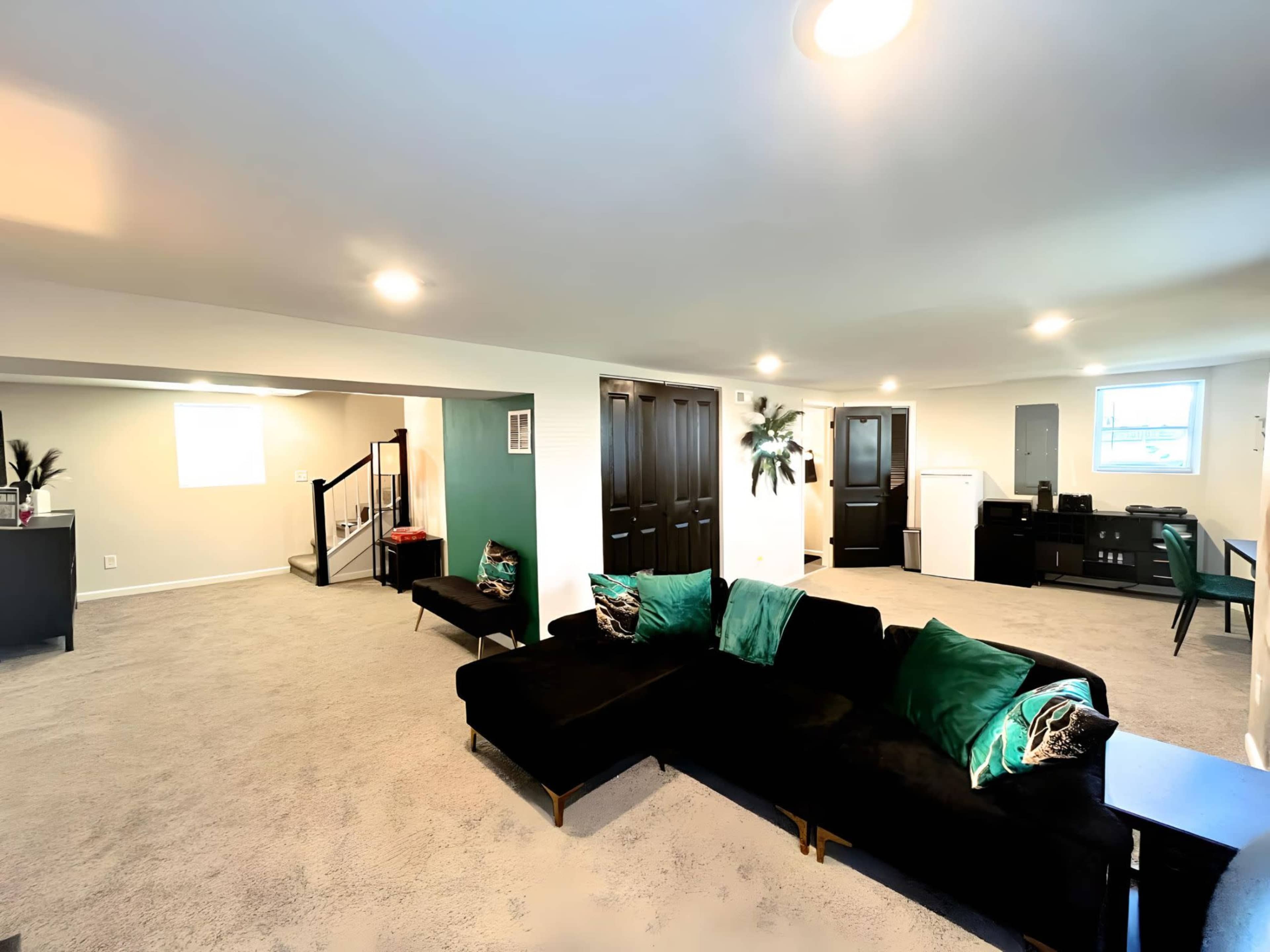 A spacious, carpeted basement with a black sectional sofa, a small desk, and multiple doors leading to separate rooms.