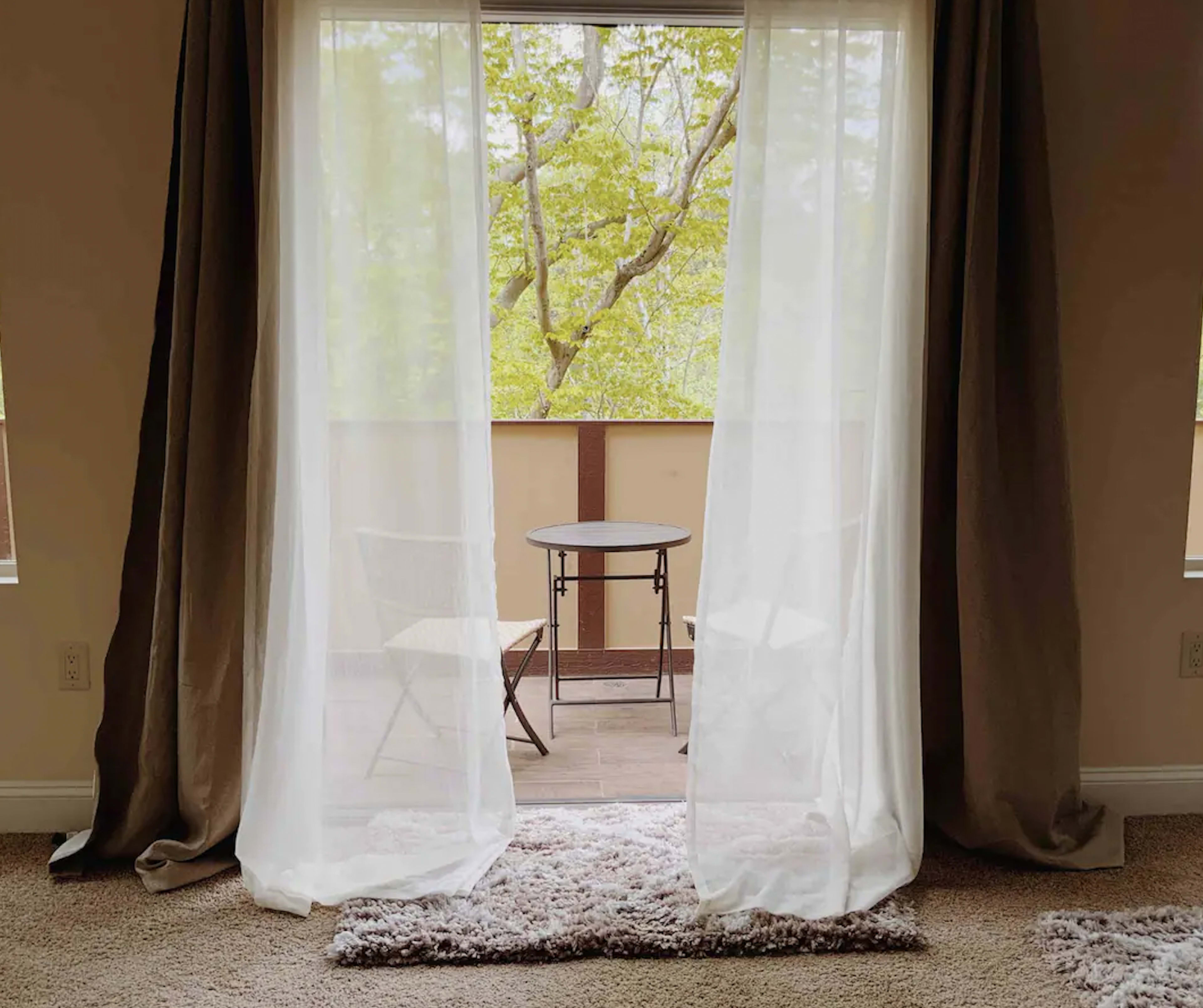 A pair of sheer curtains frame a view of a balcony with two chairs and a small table overlooking greenery.