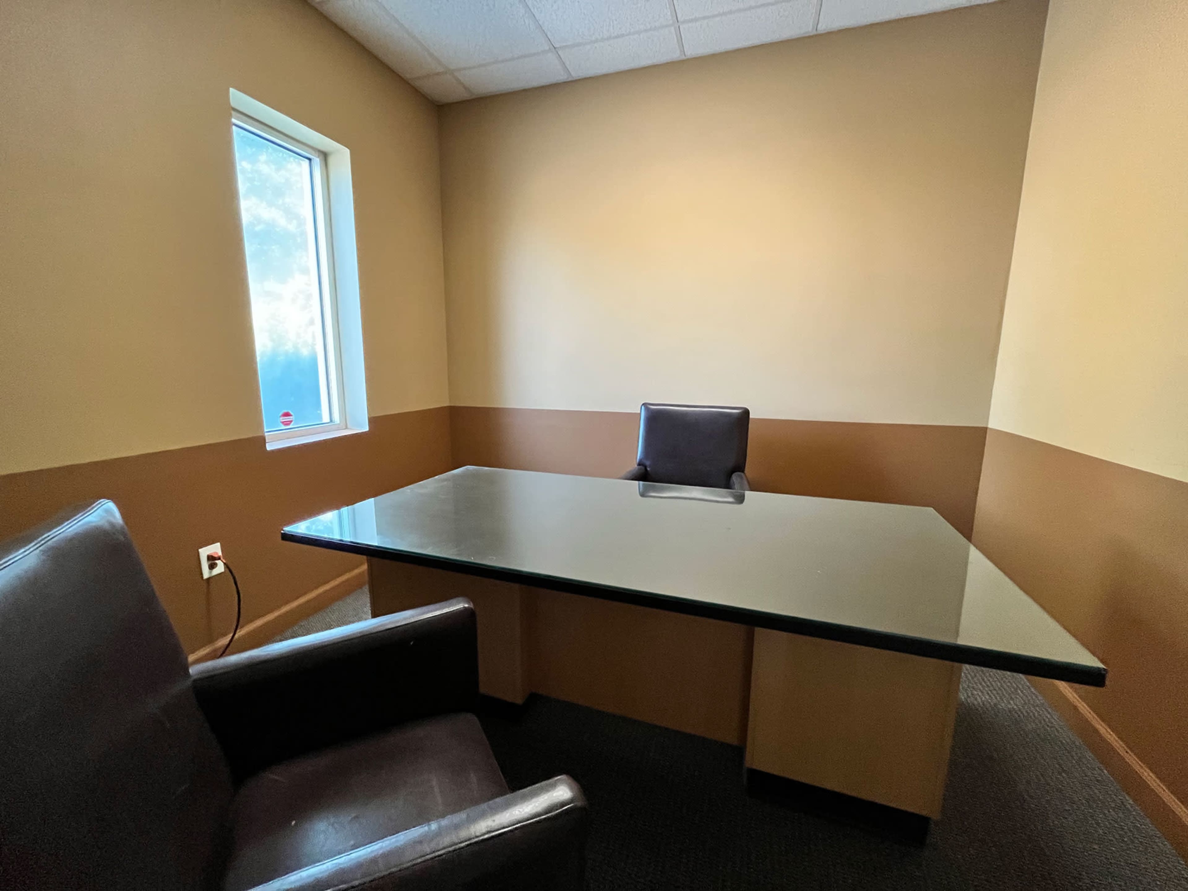 An empty office space with a glass-top desk and a single black chair facing a window, alongside two brown chairs.