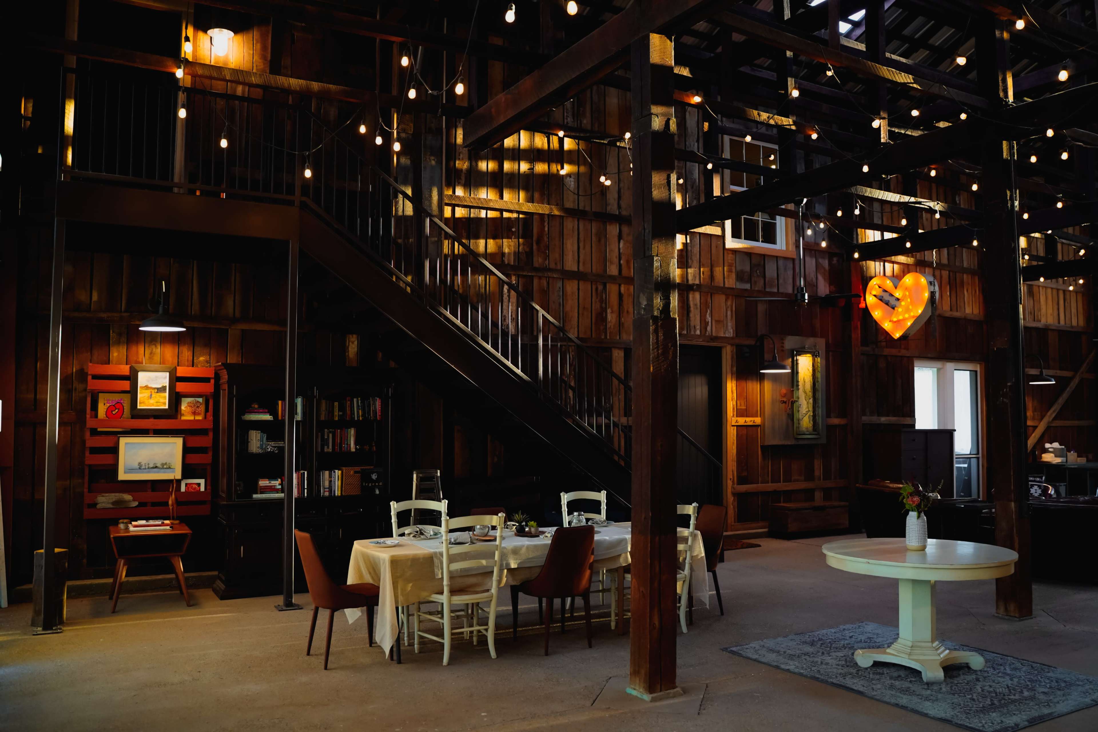 The interior of a rustic, wooden barn features a dining area with a table and chairs beneath string lights and a heart-shaped decor piece.