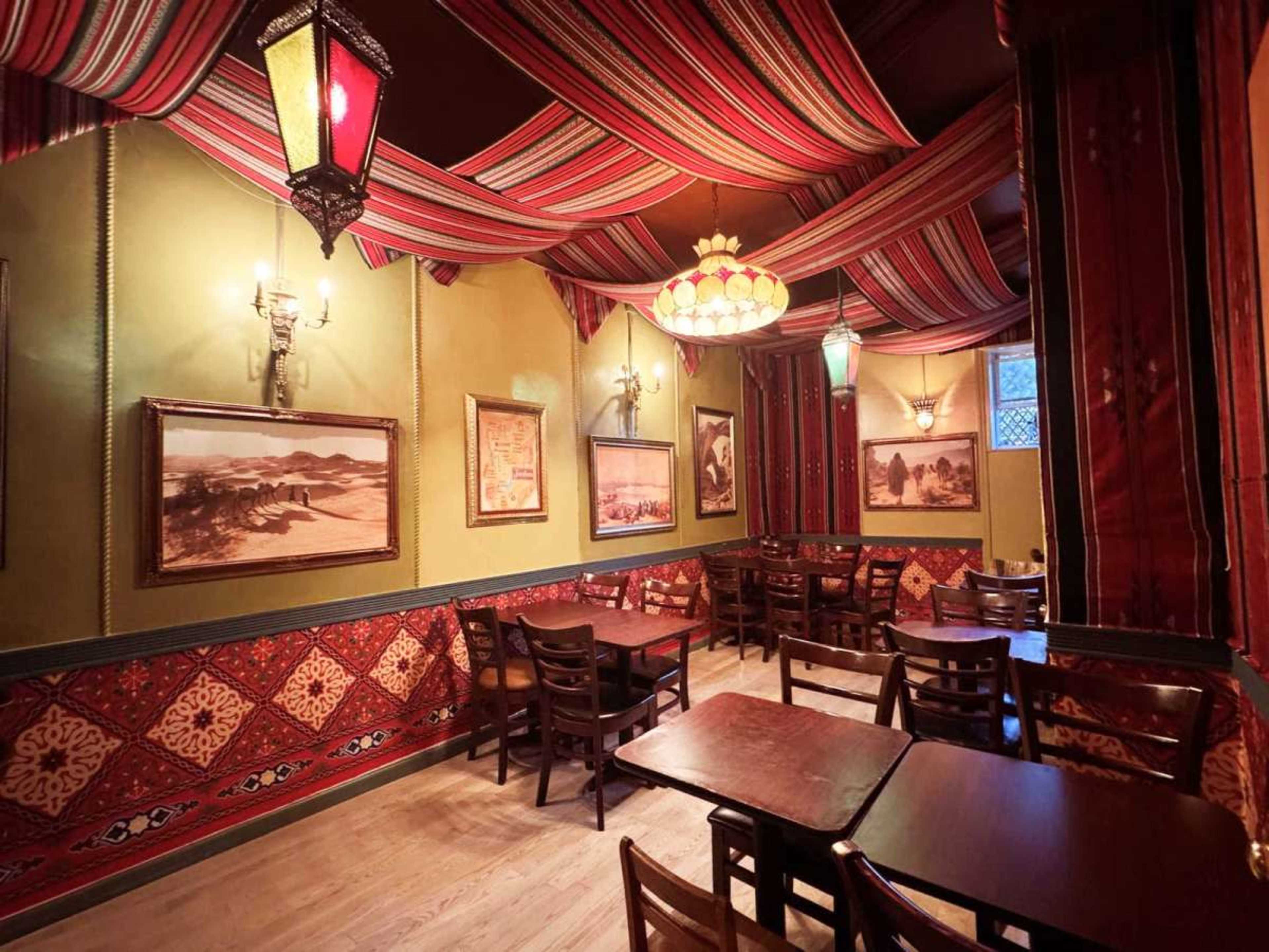 The interior of a restaurant features striped fabric draped from the ceiling, pairs of lanterns, and wooden tables arranged against walls adorned with framed photographs.