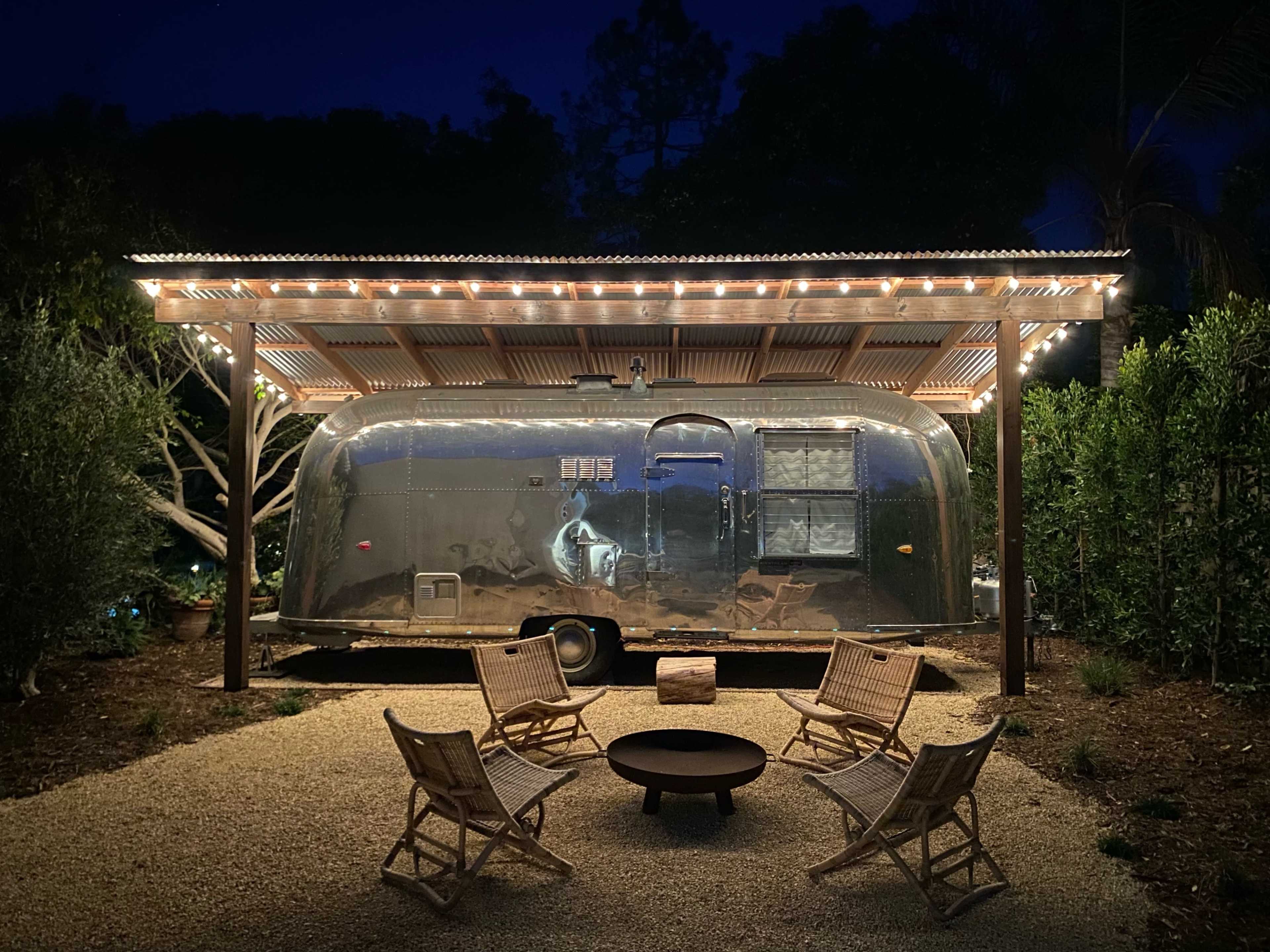 An Airstream trailer is parked under a lit wooden pergola surrounded by chairs and a fire pit at night.