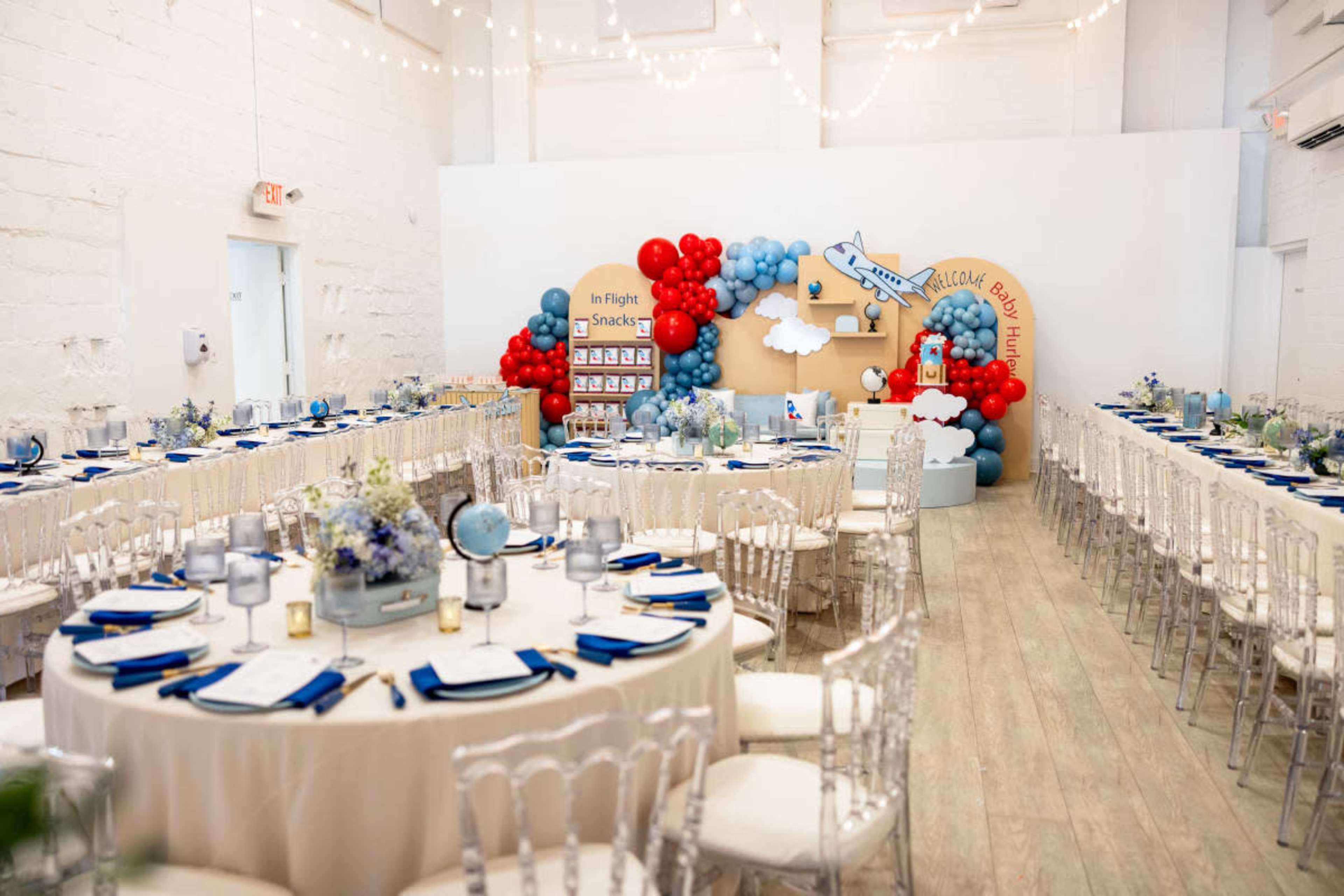 A decorated banquet hall featuring round tables with white tablecloths, blue napkins, and a backdrop of balloons and airplane-themed decor.