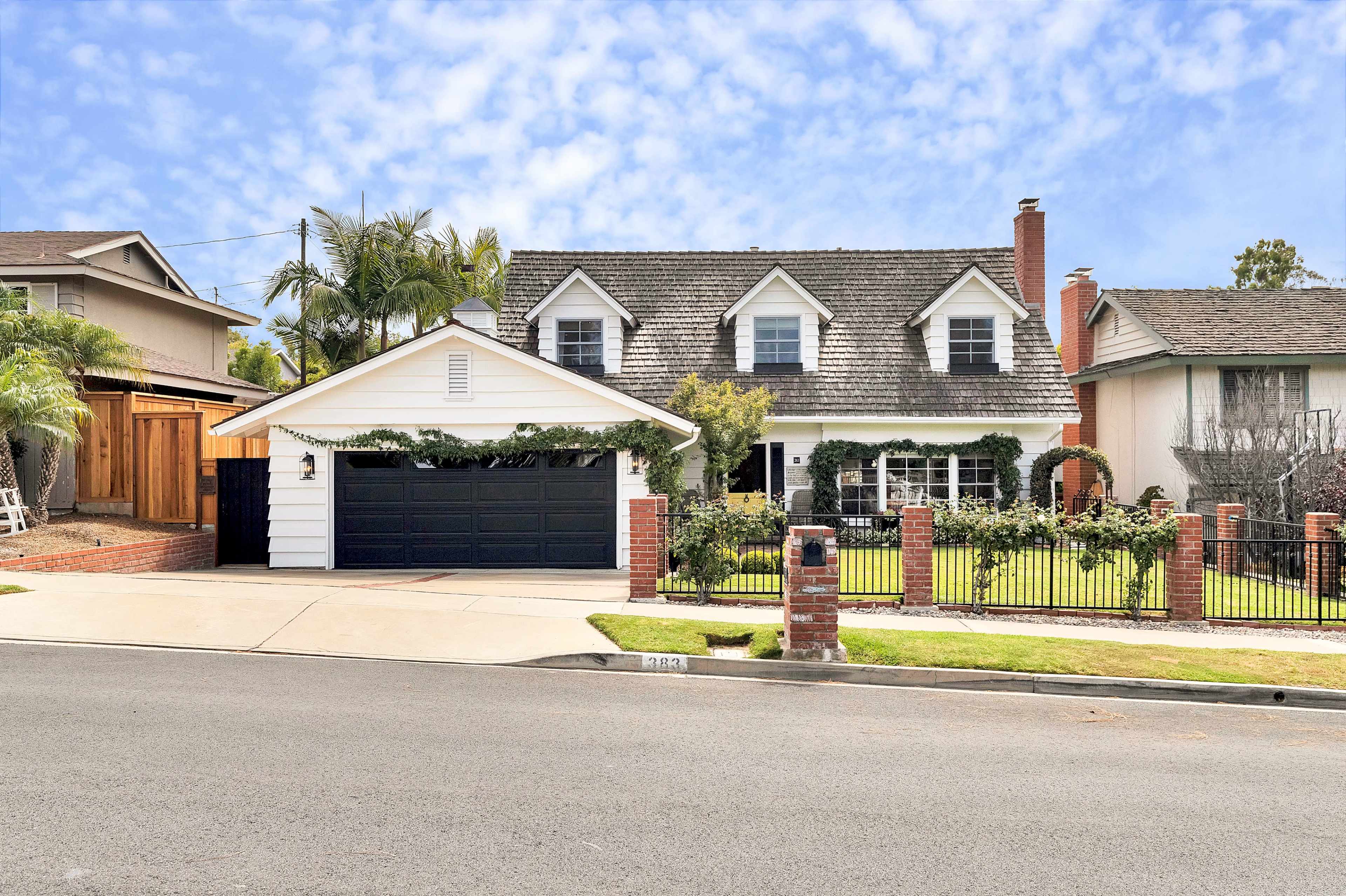 A two-story house with a steep roof, front porch, and a black garage door is surrounded by a landscaped yard and fence on a quiet street.