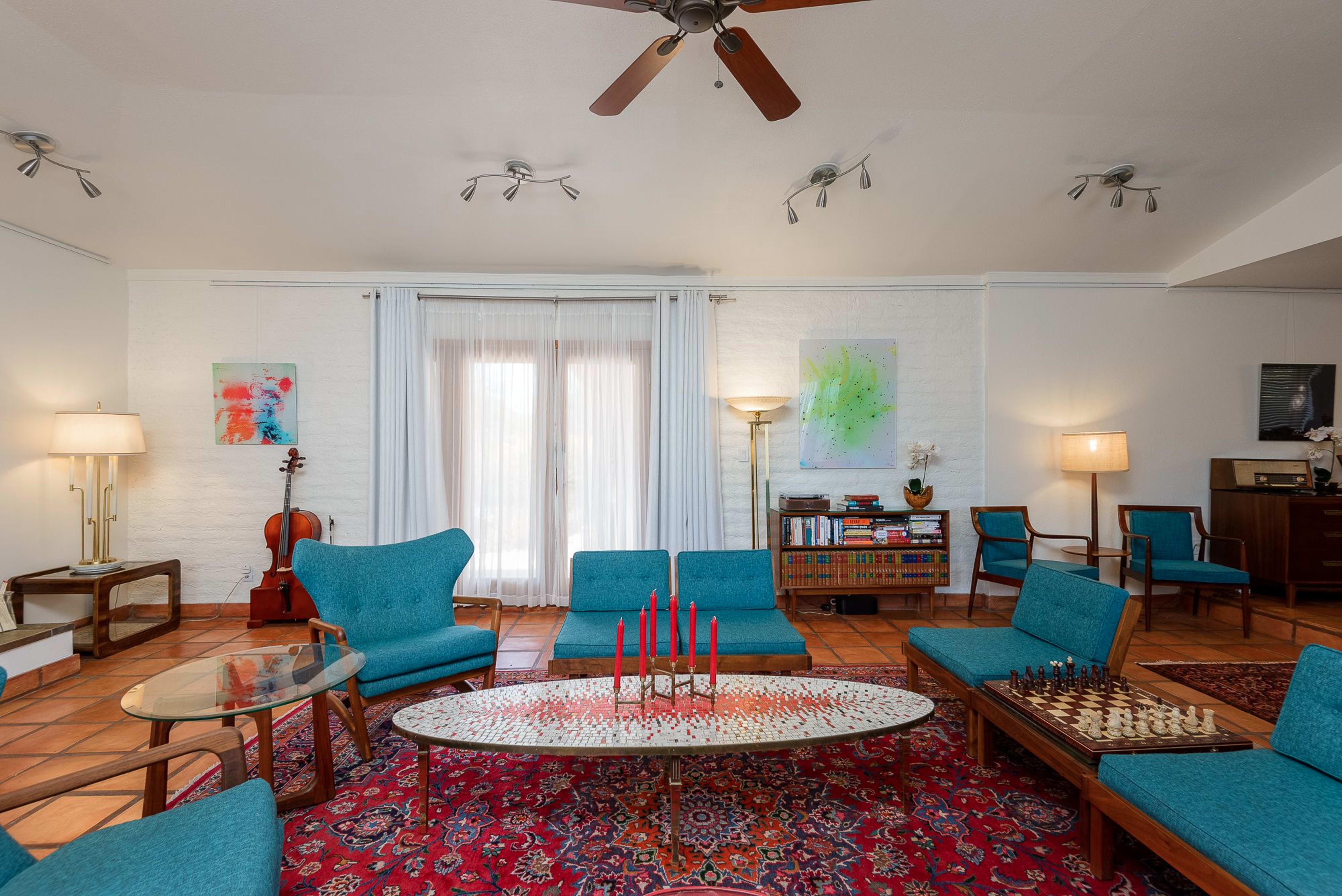 The room features a seating area with blue chairs arranged around a patterned rug, a coffee table with red candles, and a wooden chess set, alongside a window with sheer white curtains.