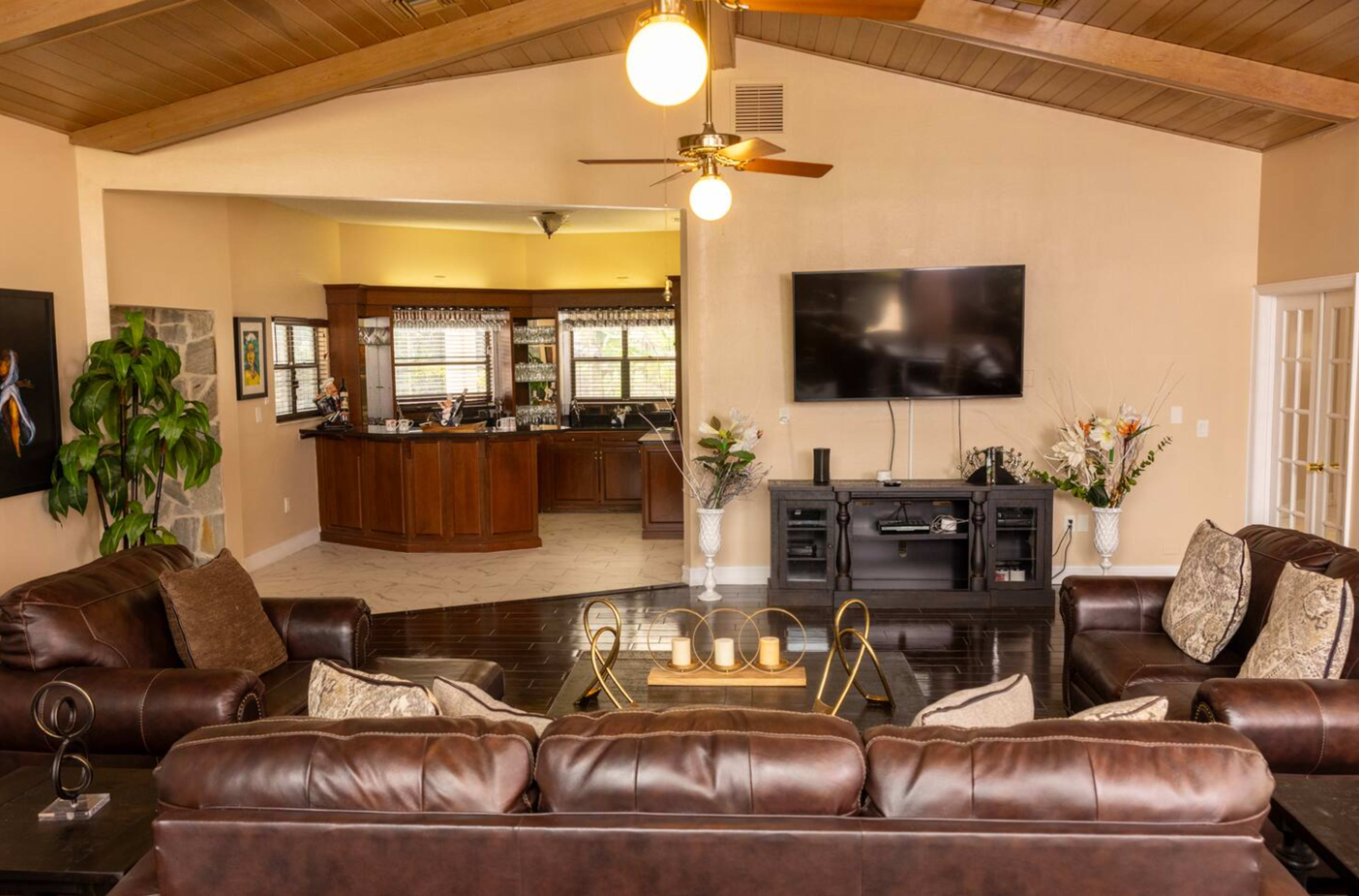 The image shows a spacious living room with a brown leather sectional sofa, a coffee table, and a large television mounted on the wall, with a view into a kitchen area in the background.