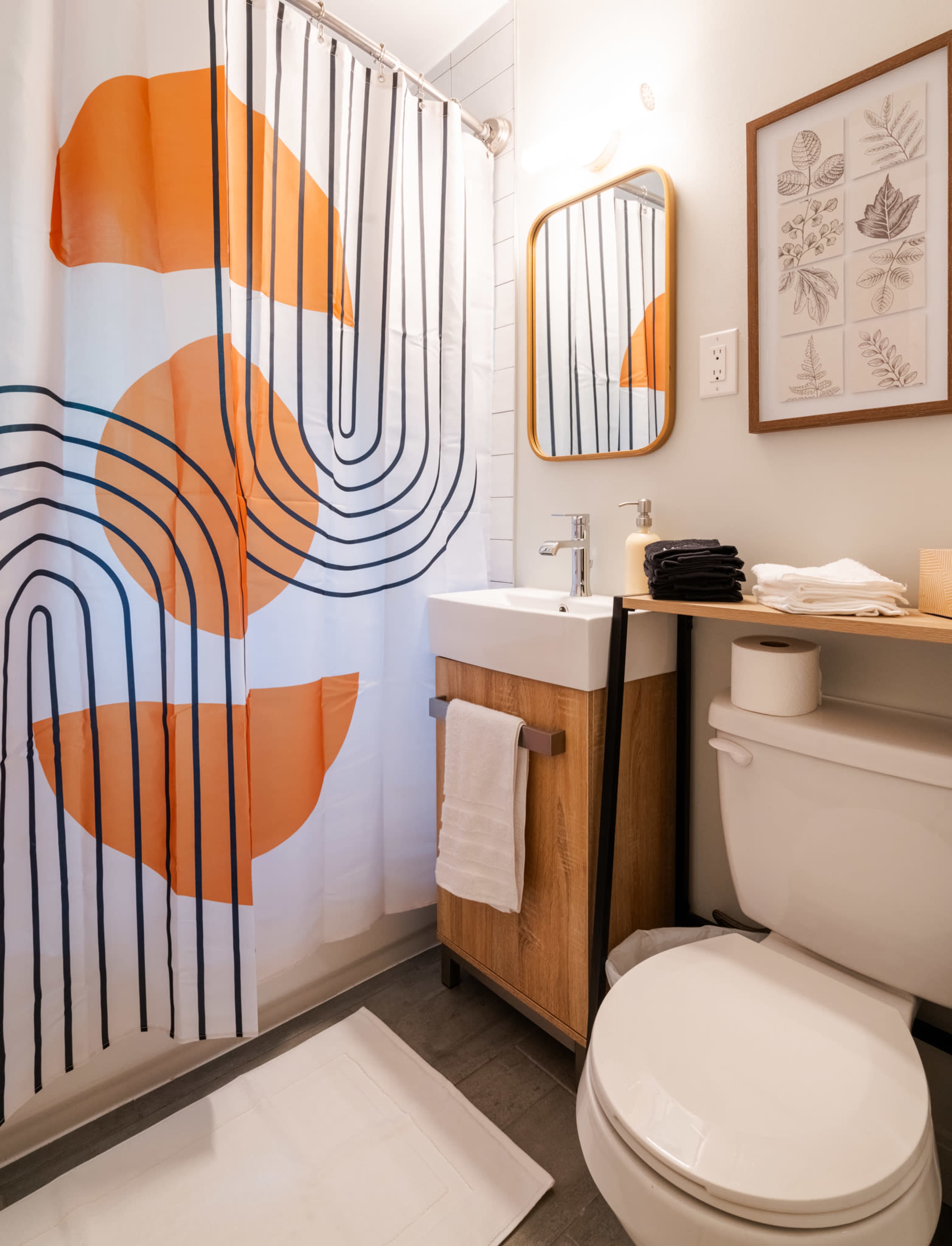 A modern bathroom featuring a colorful shower curtain with orange and black designs, a wooden vanity with a sink, a mirror, and neatly arranged towels and toiletries.