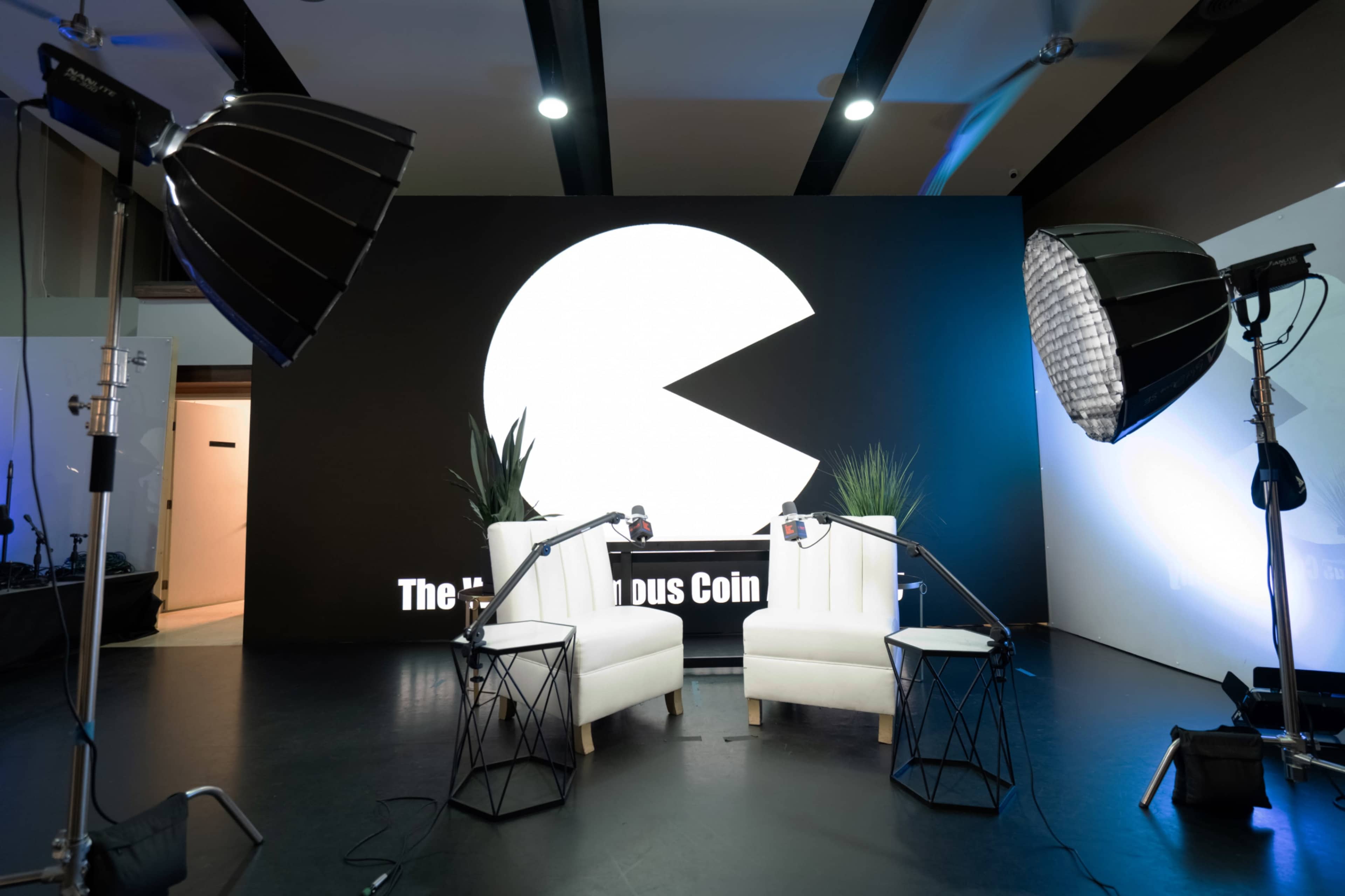 The image shows two white chairs facing each other in a studio setting, with large softbox lights positioned on either side and a prominent circular graphic resembling Pac-Man on the backdrop.