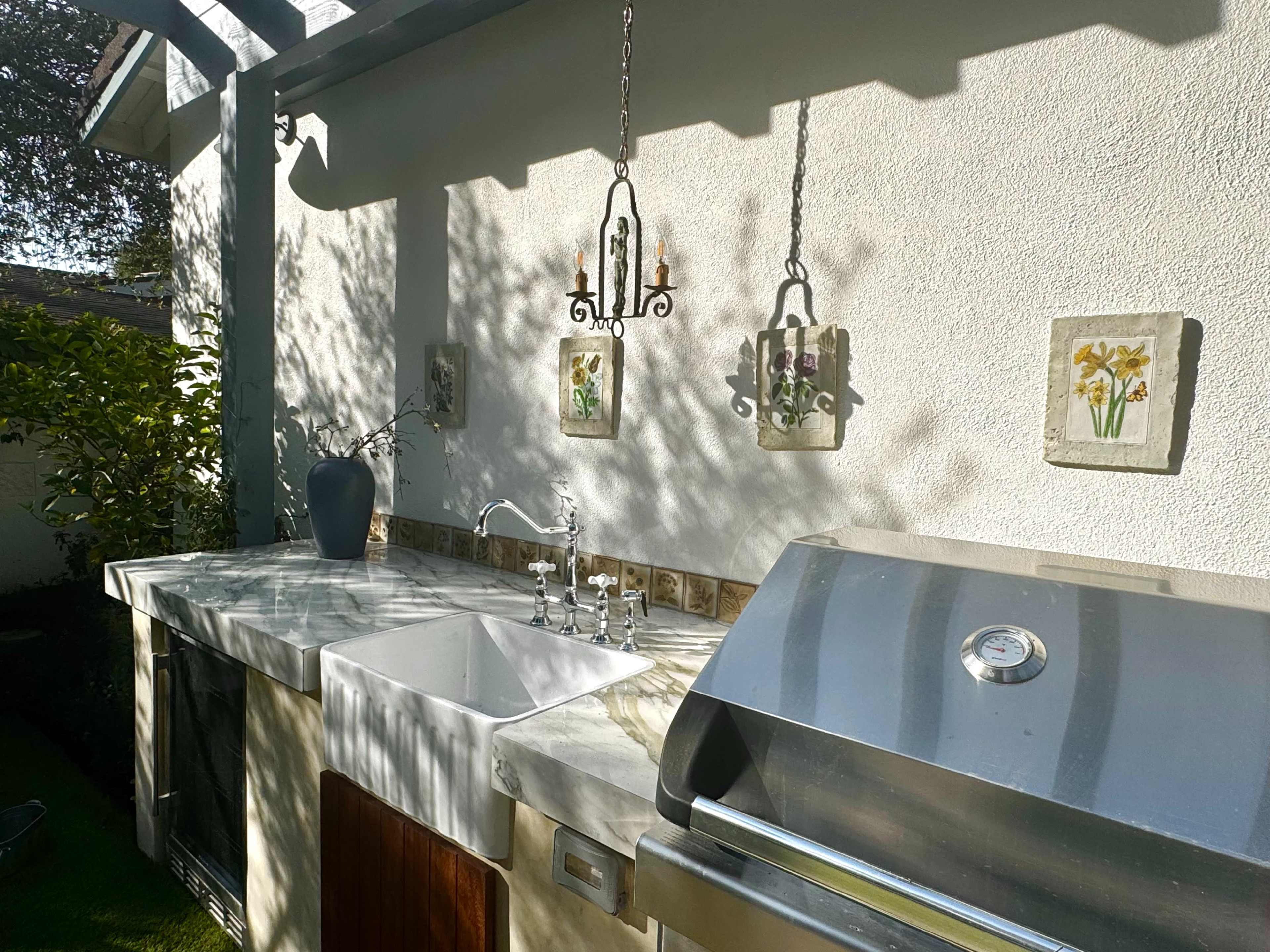 The image shows an outdoor kitchen area with a marble countertop, a stainless steel grill, and decorative wall art featuring flowers.
