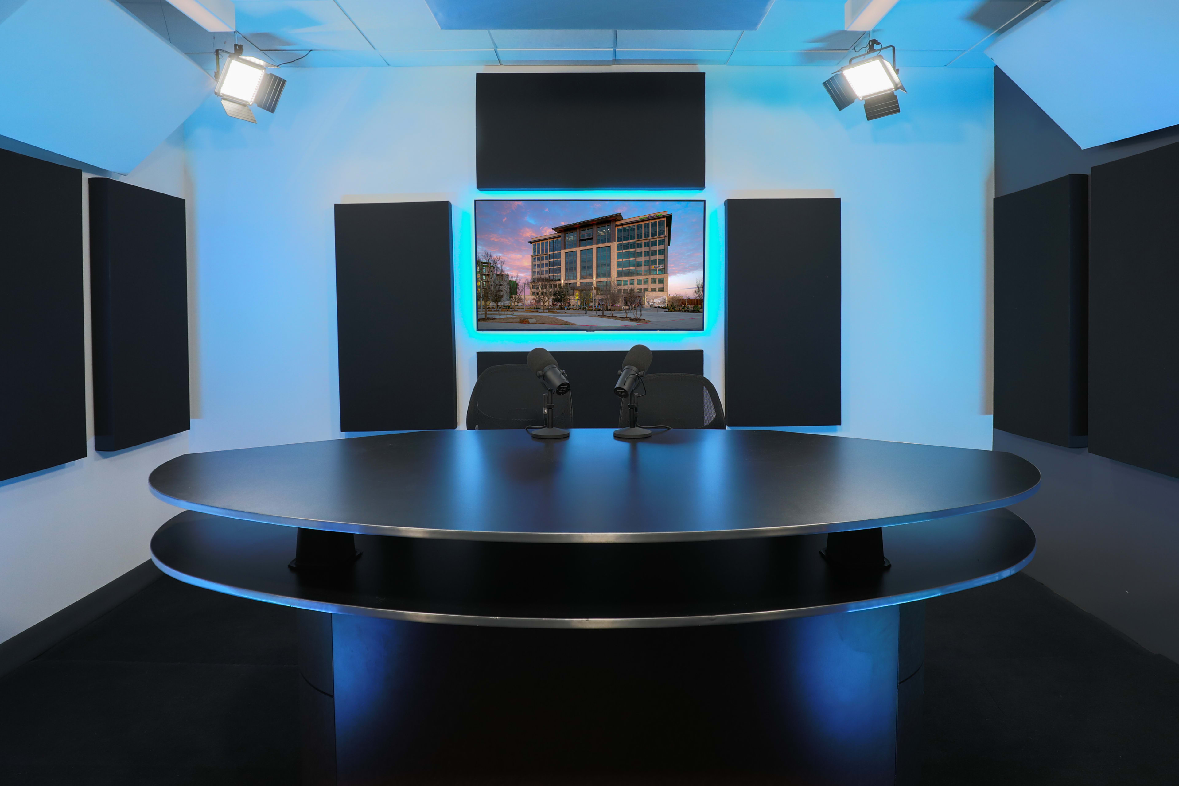 A modern broadcast studio features a curved black table with two microphones and a large screen displaying a building construction image in the background.