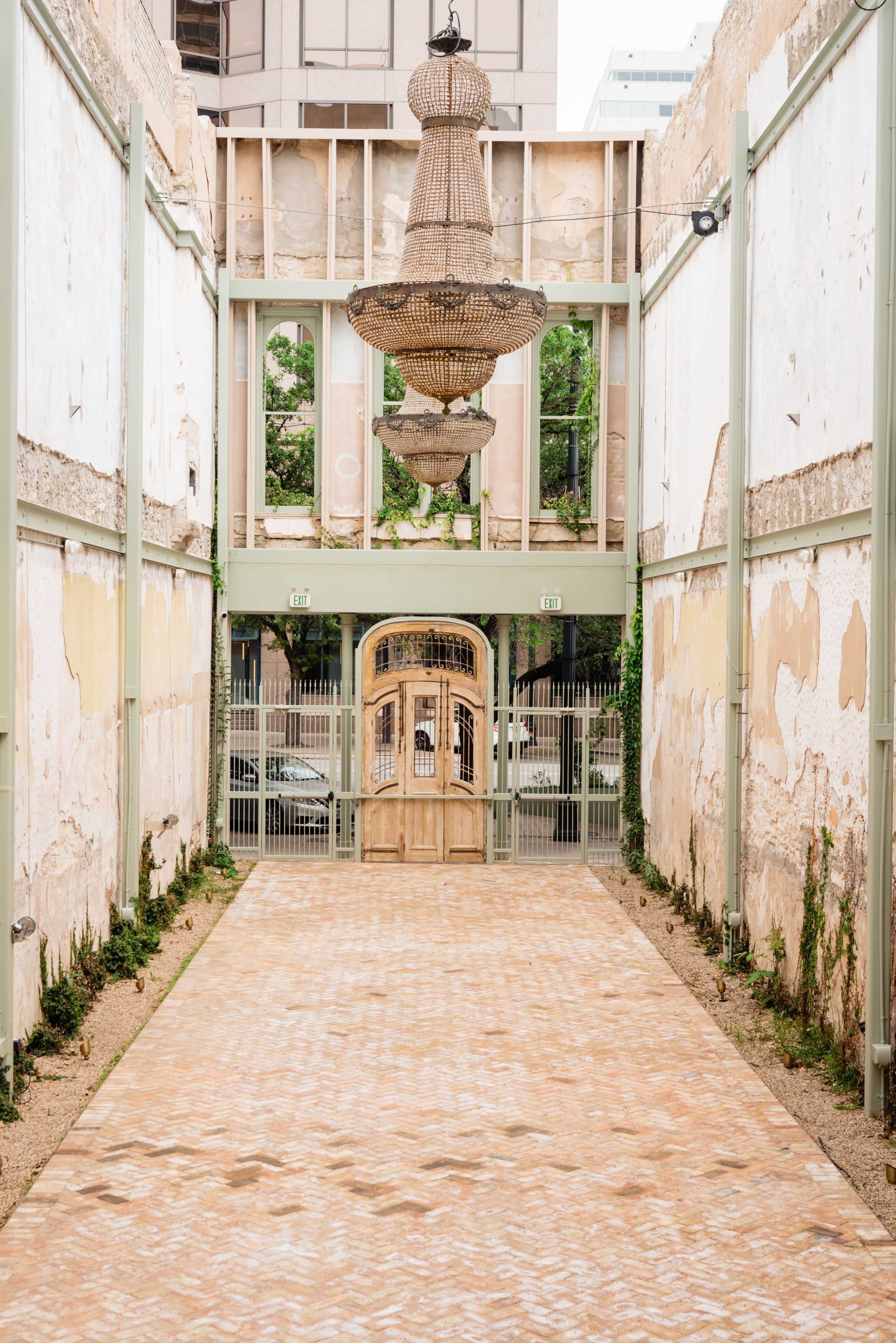 The image shows a narrow, weathered corridor with a decorative chandelier hanging from the ceiling and an ornate wooden door at the end, flanked by crumbling walls.