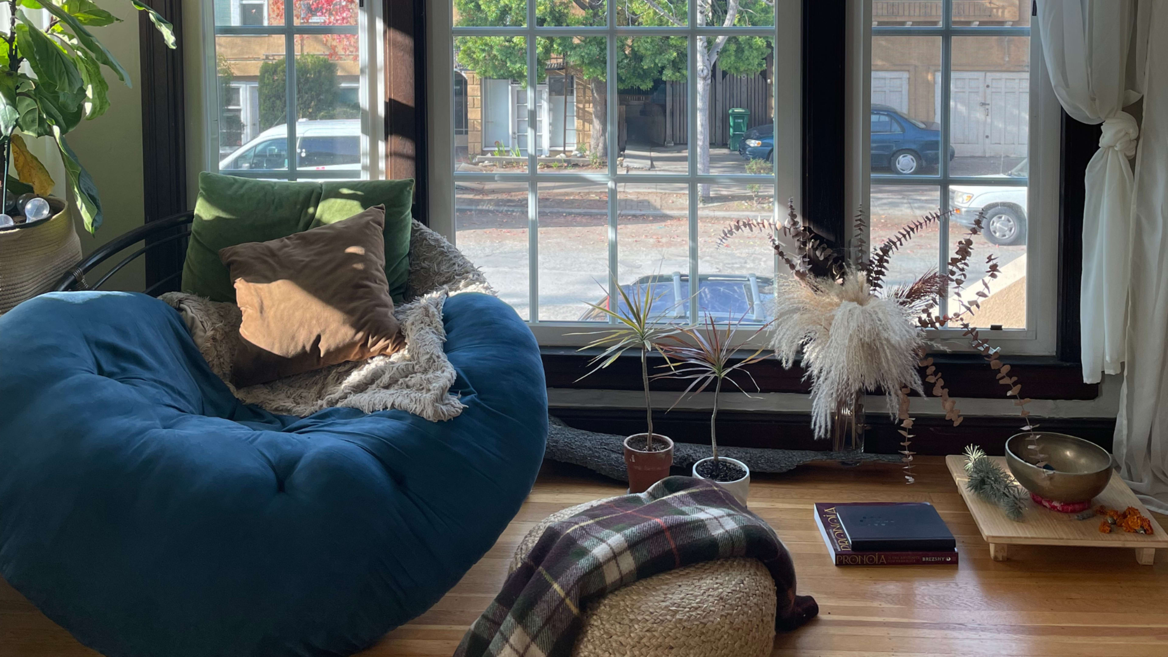 A cozy corner with a large blue bean bag chair next to potted plants and a window overlooking a street.