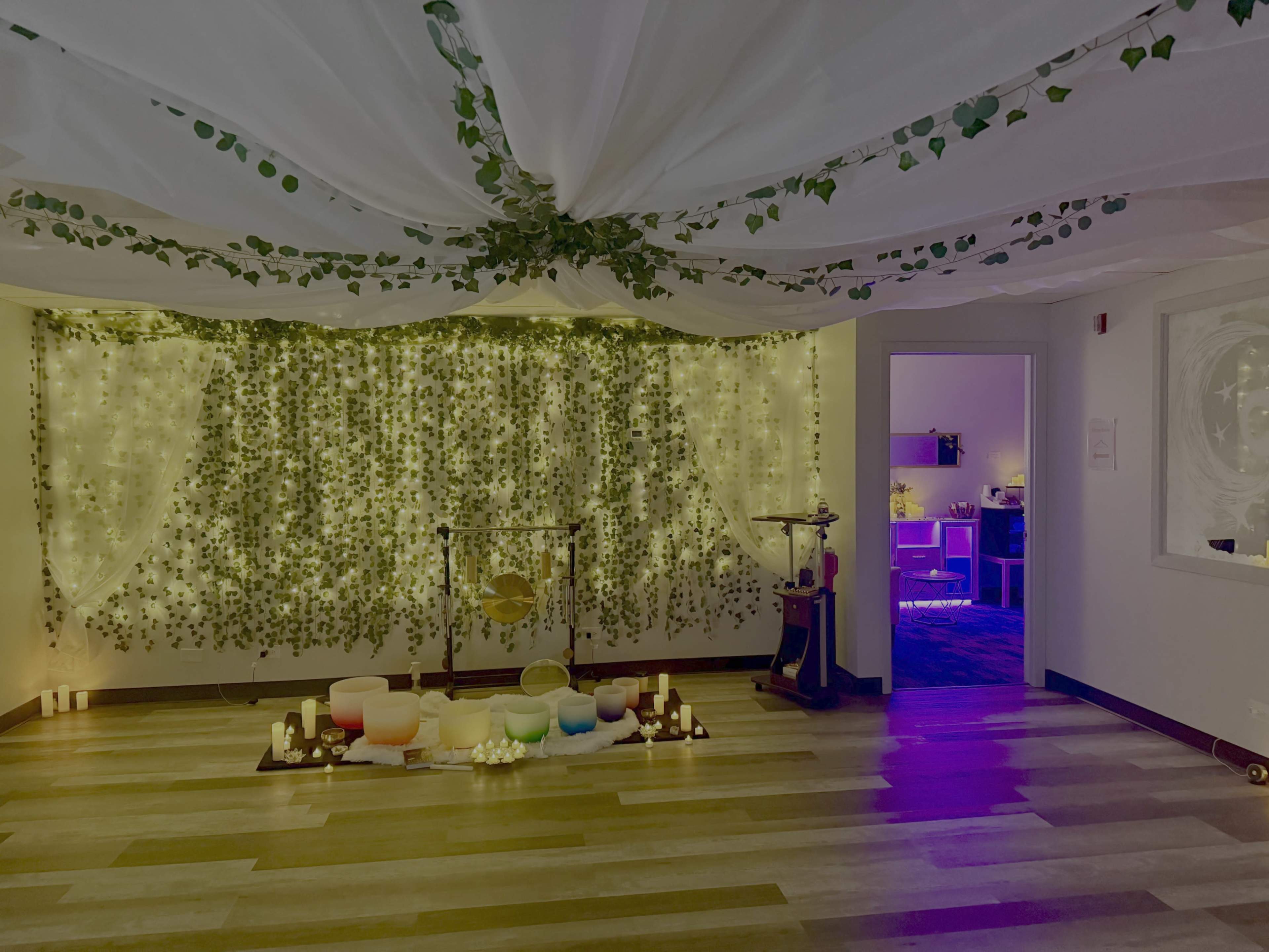 The image shows a dimly lit room adorned with string lights and green foliage, featuring a setup of crystal bowls arranged on a mat, with candles placed around them.