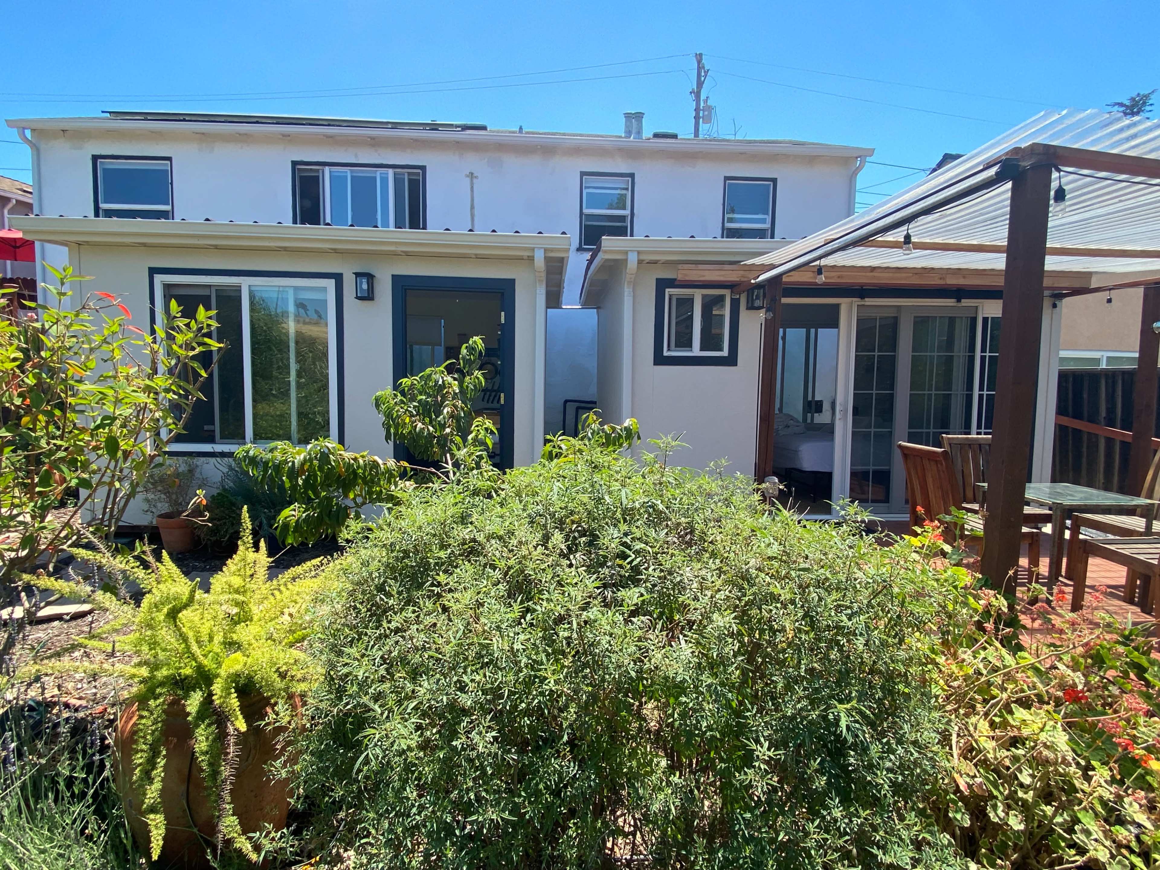 A residential backyard featuring two adjacent buildings surrounded by greenery and a patio area.