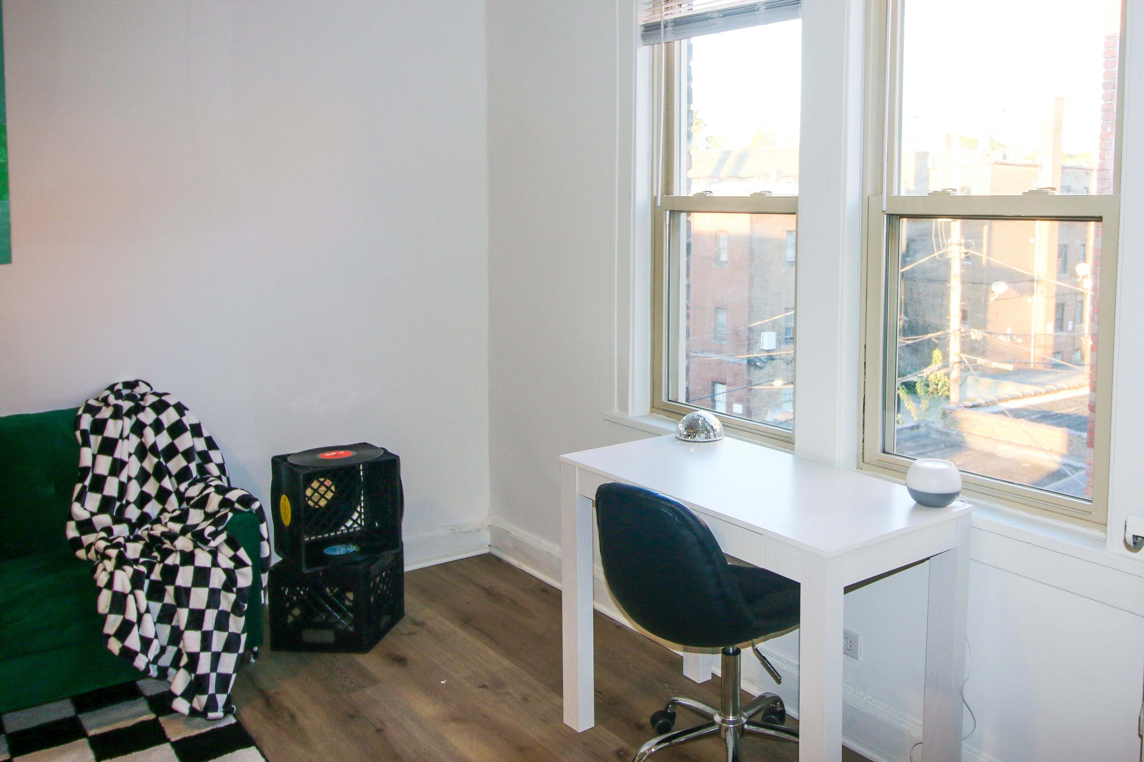 A simple room featuring a green couch with a black and white checkered blanket, a white desk with a chair, and two windows revealing an outdoor view.