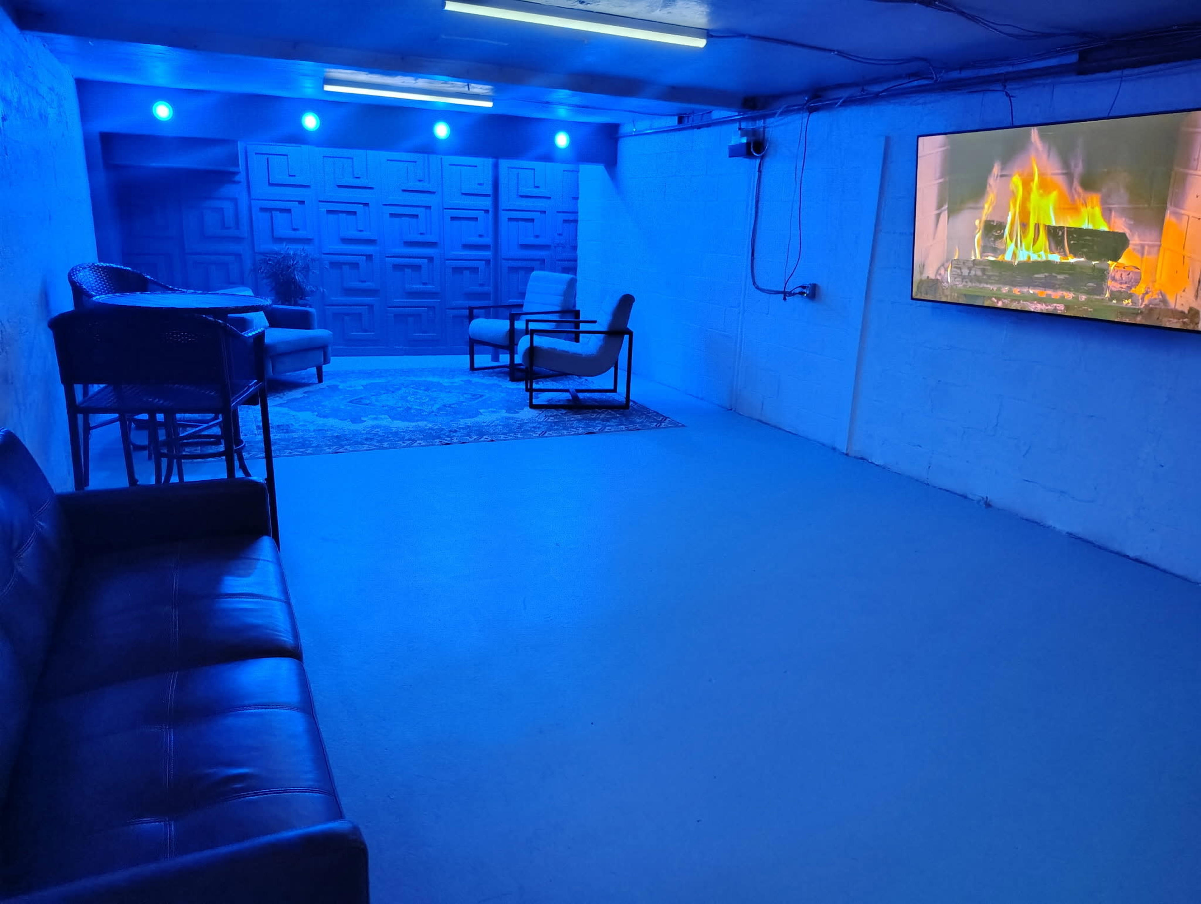 The image shows a blue-lit basement room with a couch, two chairs, a small table, and a large screen displaying a fireplace.