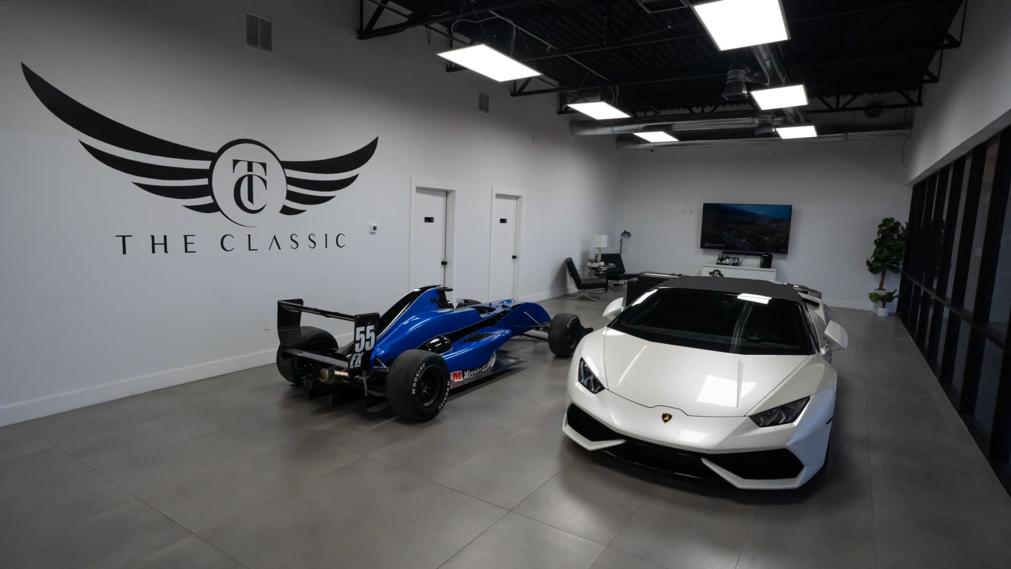 A modern showroom featuring a blue racing car and a white sports car, with a large wall logo that reads "THE CLASSIC."