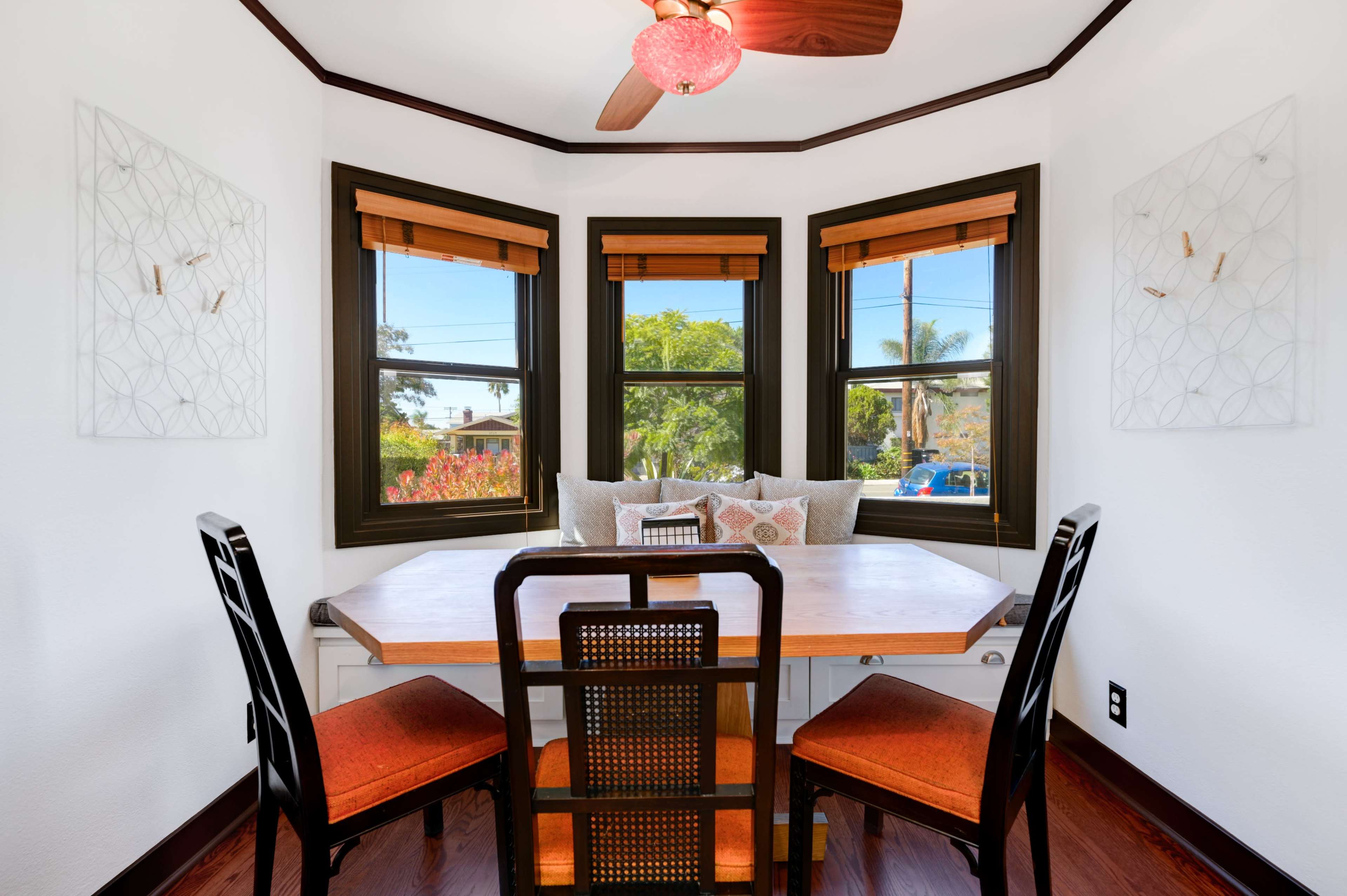 A small dining area features a wooden table and four chairs surrounded by three windows with bamboo blinds.
