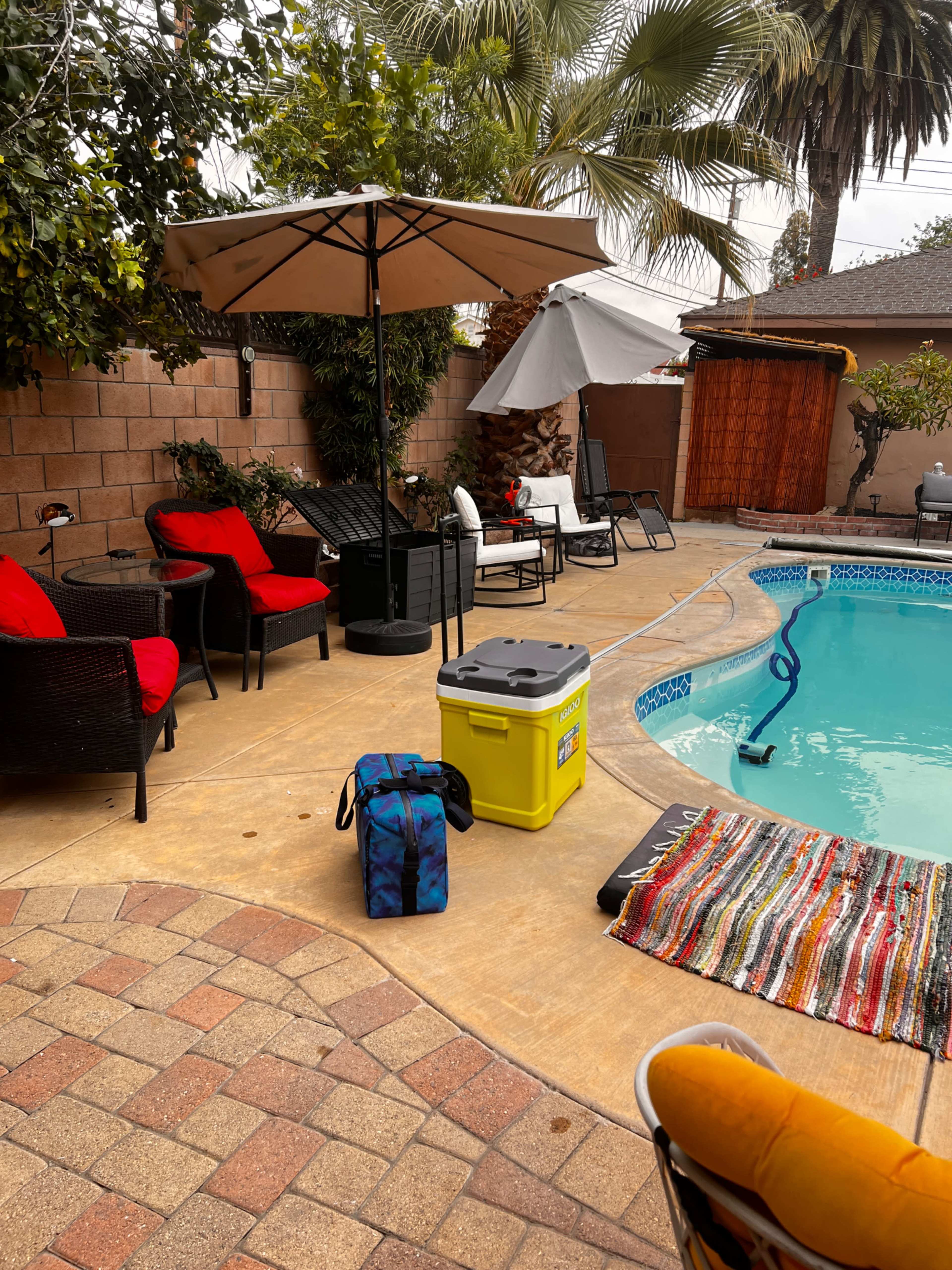 The image shows a backyard patio with a swimming pool, several seating areas, umbrellas, and a cooler placed on the ground.