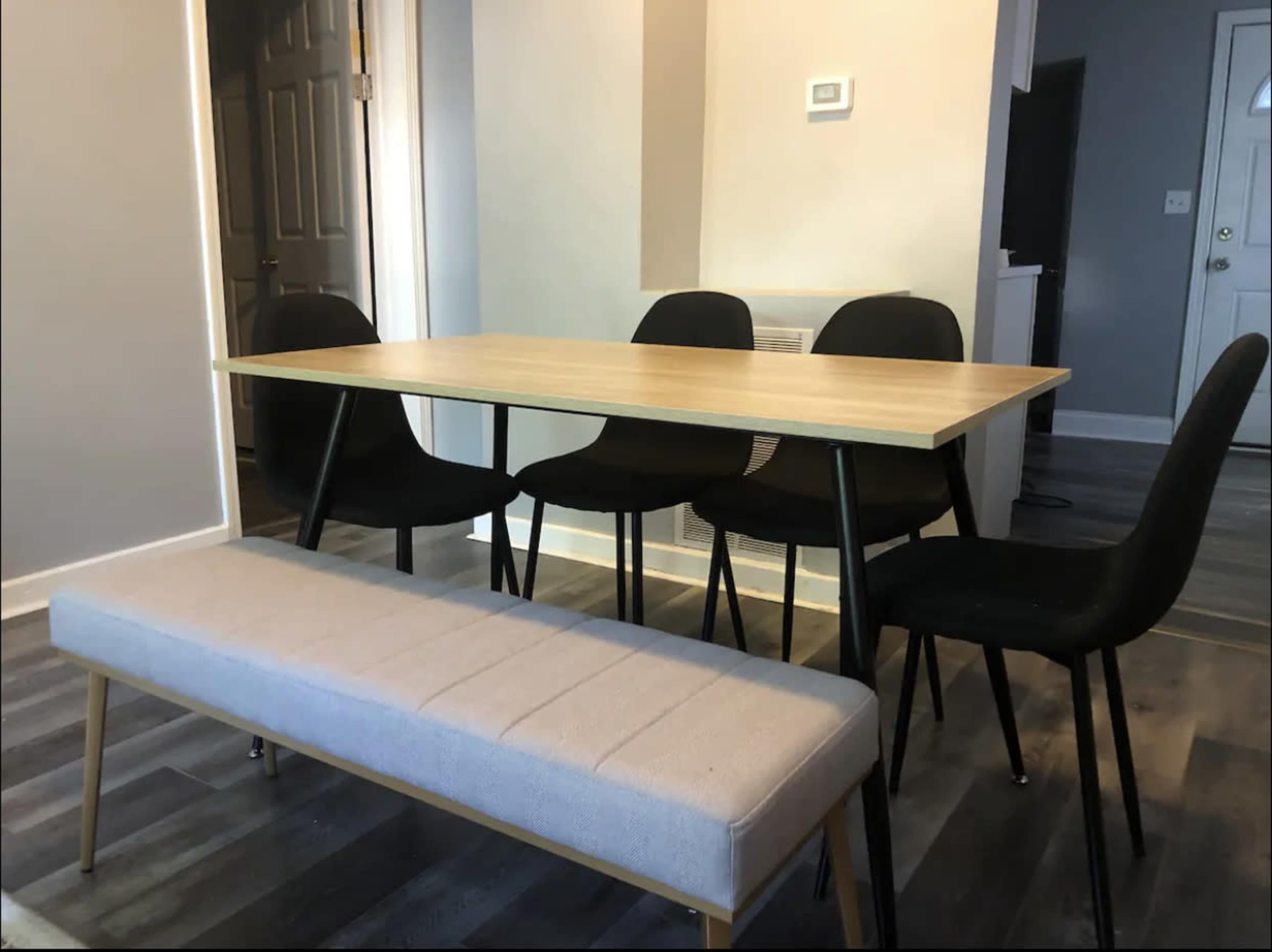 A light wood table is surrounded by four black chairs, with a gray upholstered bench positioned in front of the table.