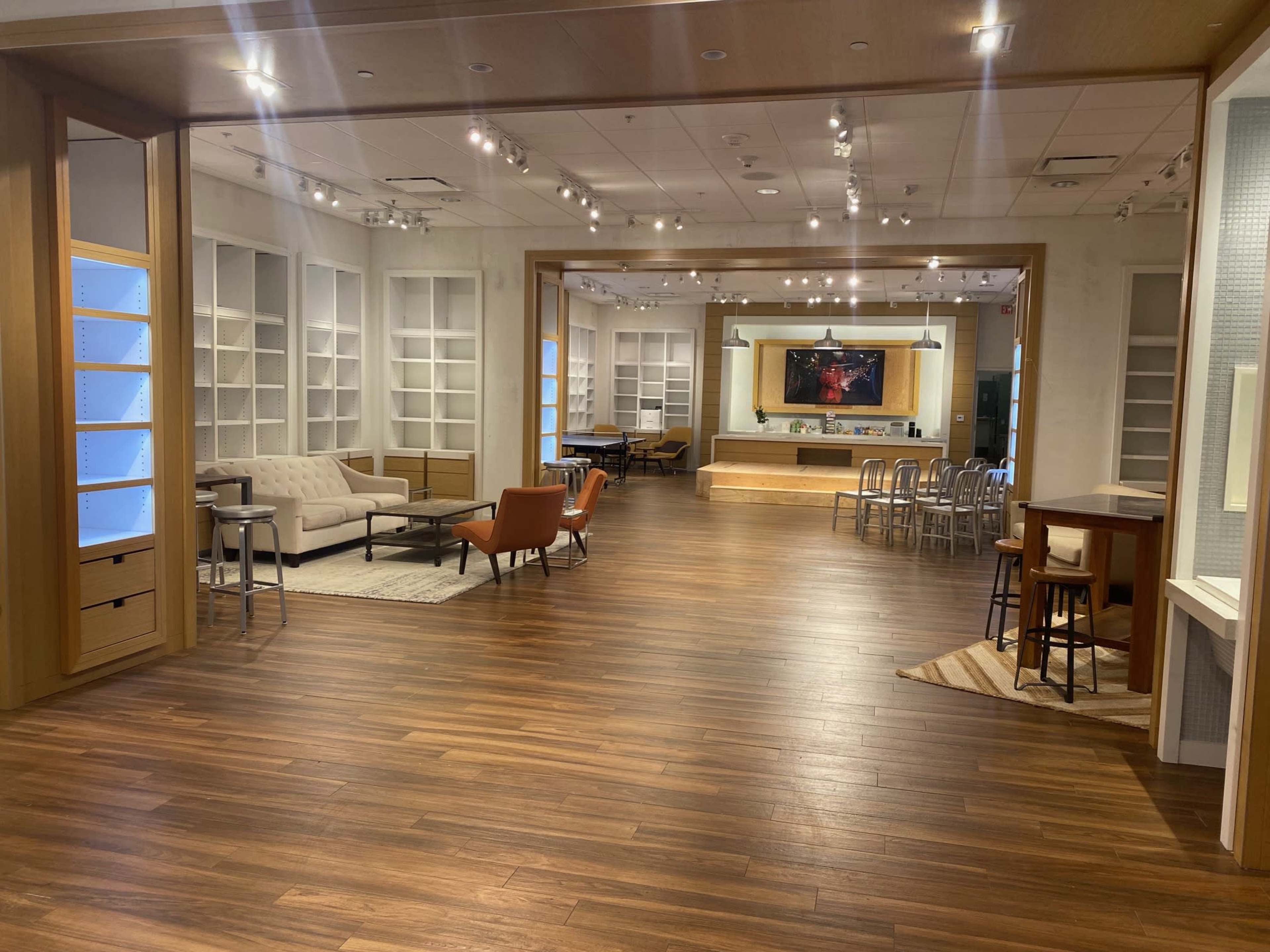 A spacious room with wooden flooring, featuring a seating area with a sofa and armchairs, a kitchen space in the background, and built-in shelves along the walls.