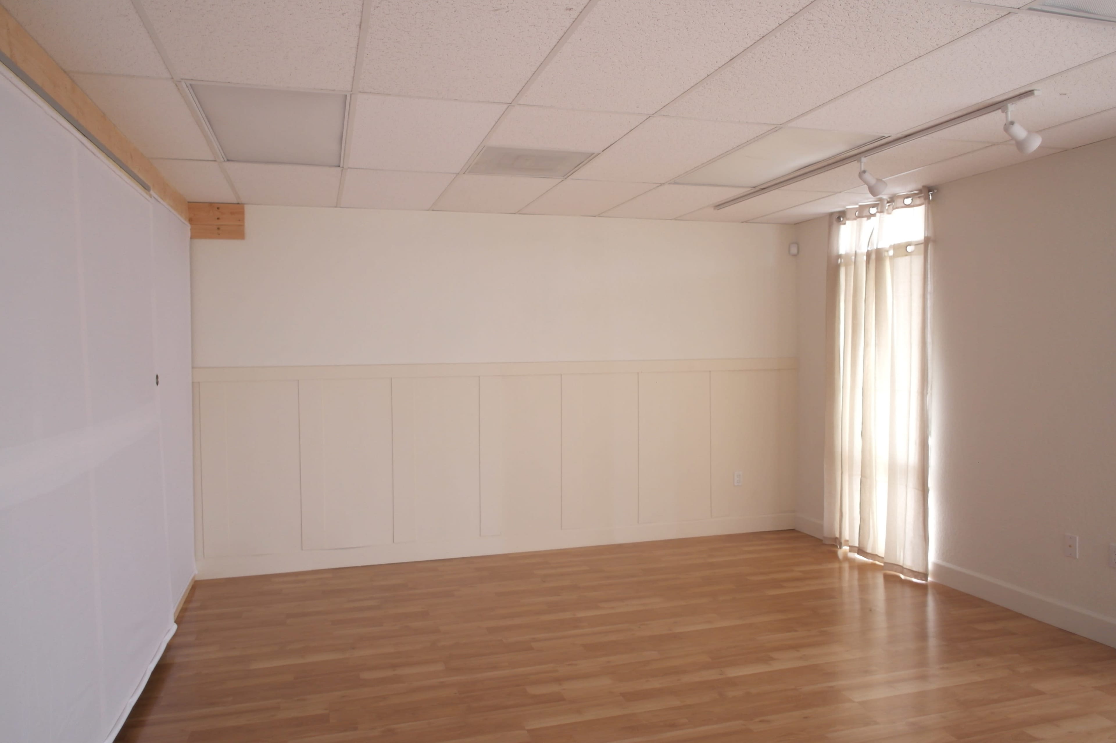 A spacious, empty room with light wooden flooring and a large window covered by sheer curtains.