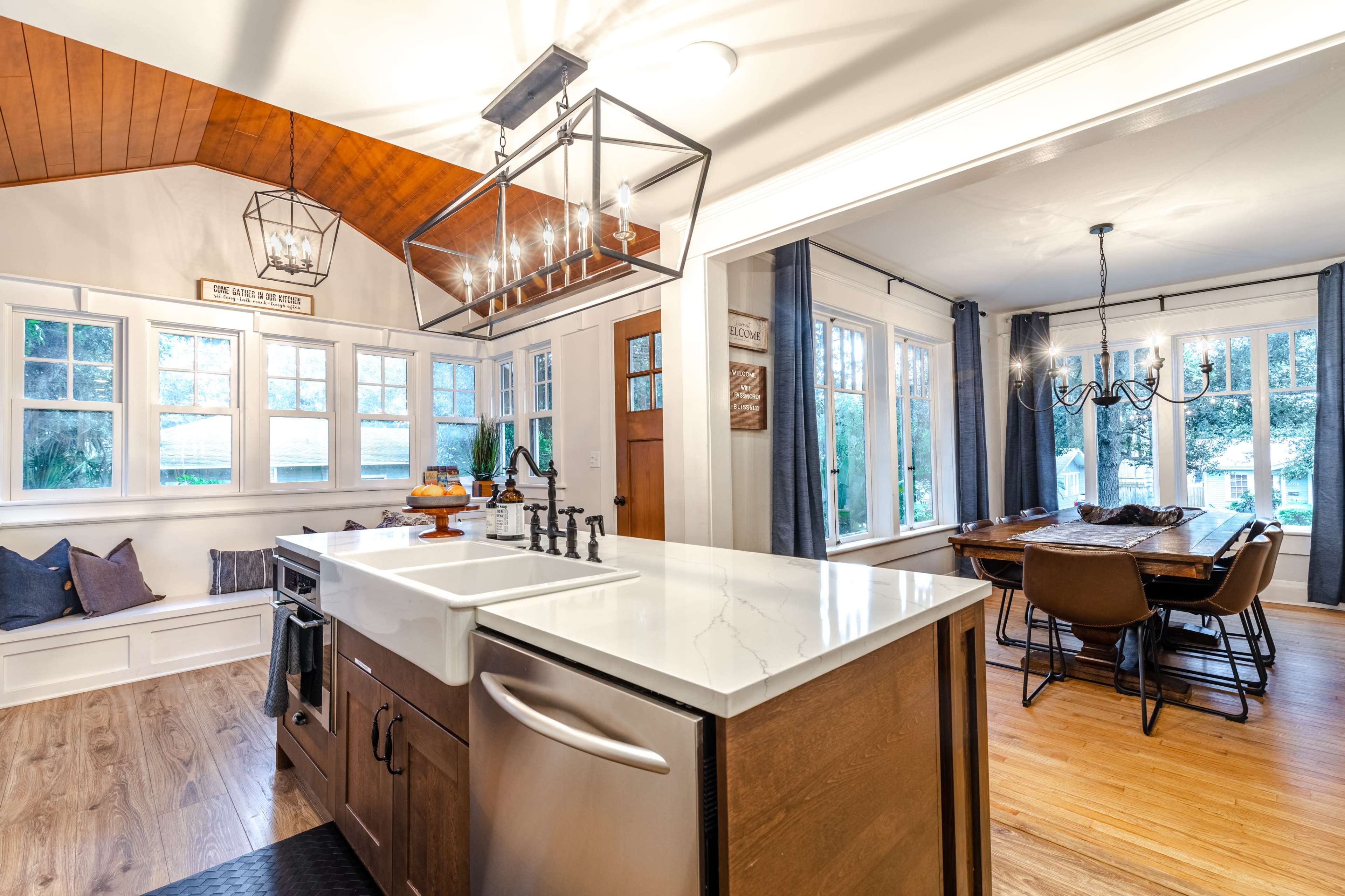 A modern kitchen with a large island, stainless steel appliances, and a dining area featuring a wooden table and six chairs, illuminated by natural light from large windows.