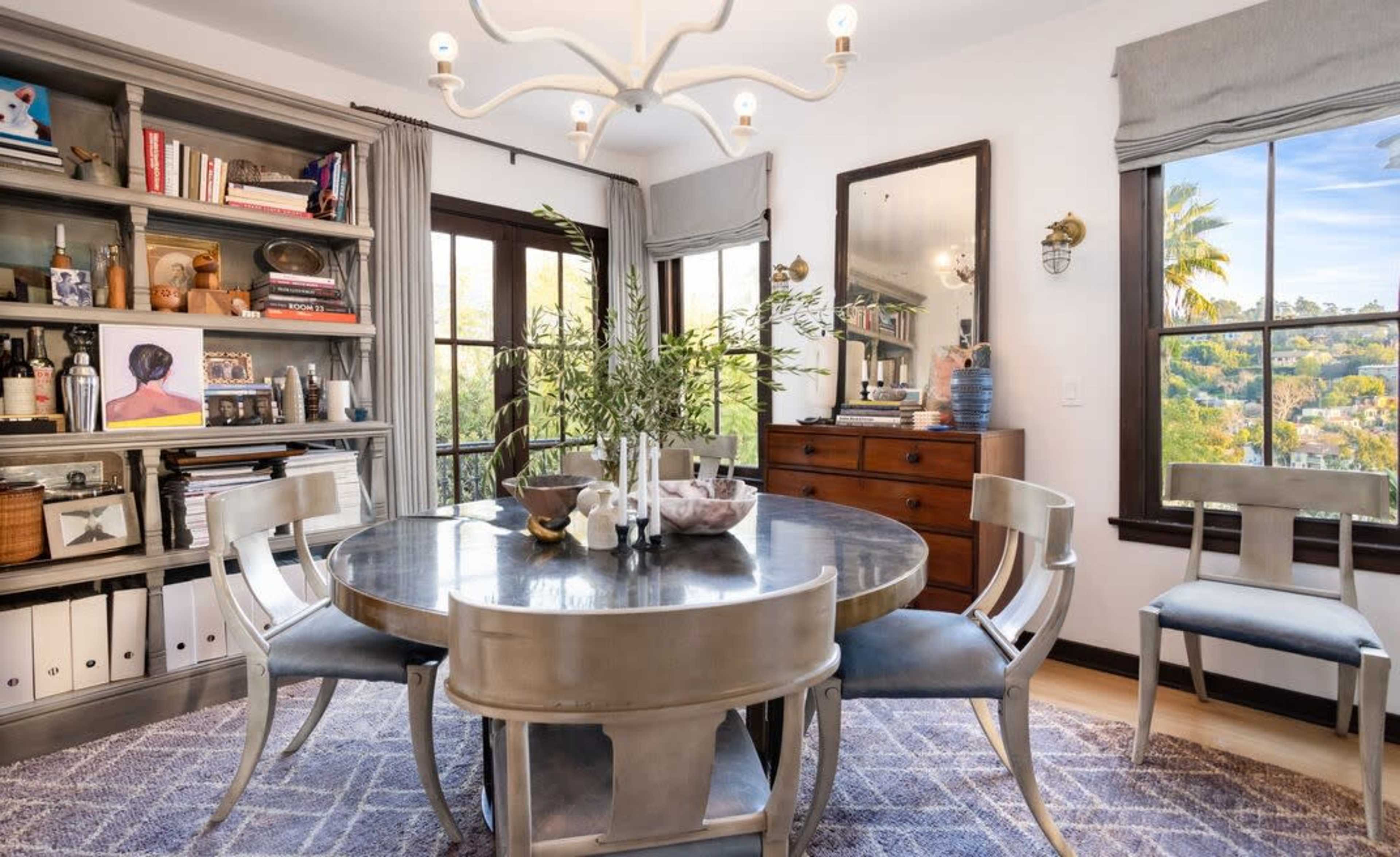 The image shows a dining area with a round table surrounded by four chairs, a bookshelf filled with books and decorative items, and large windows providing a view of the outdoors.