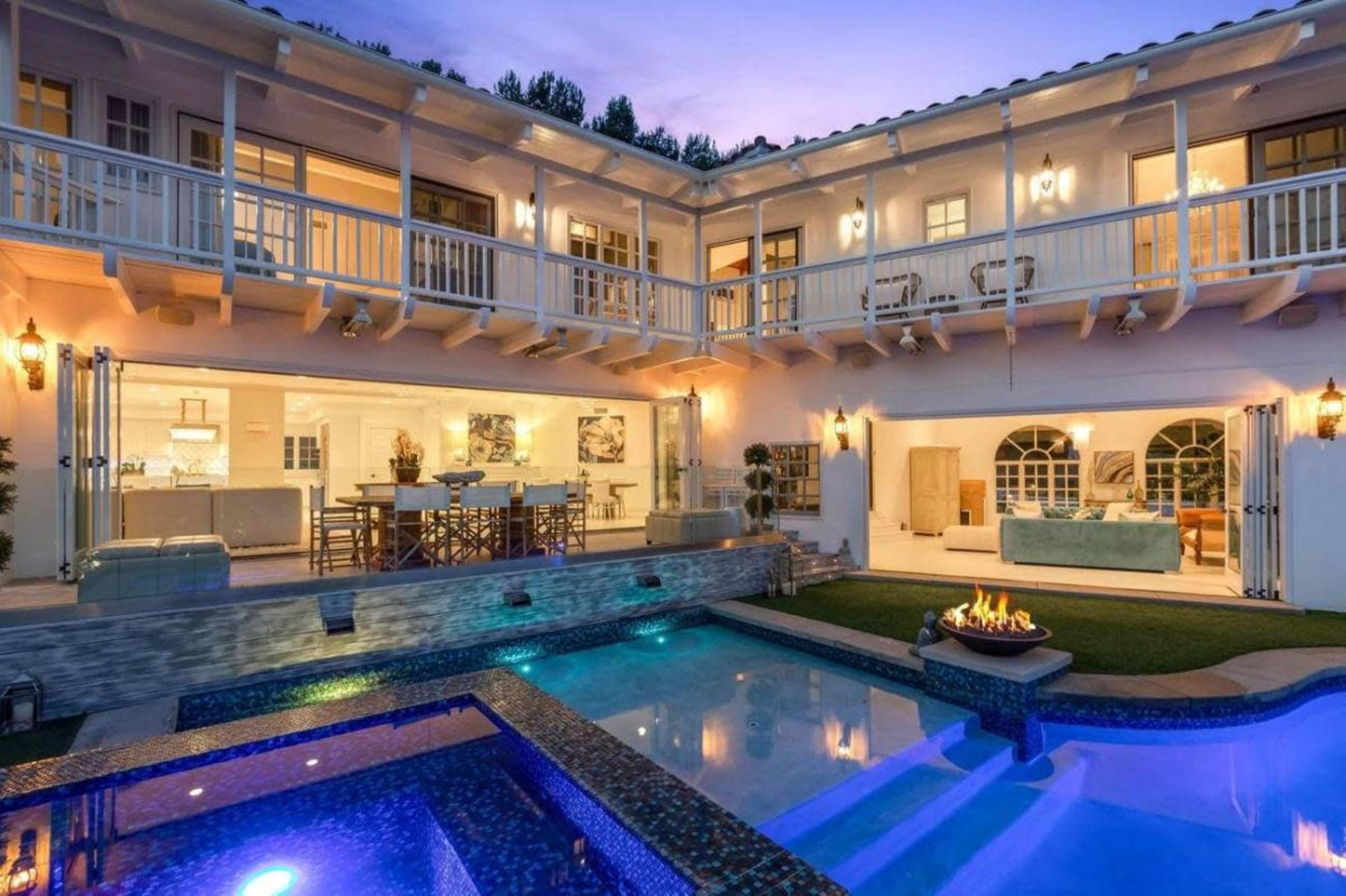 A modern two-story house features an open patio, a swimming pool, and a spa area, illuminated by evening lights.