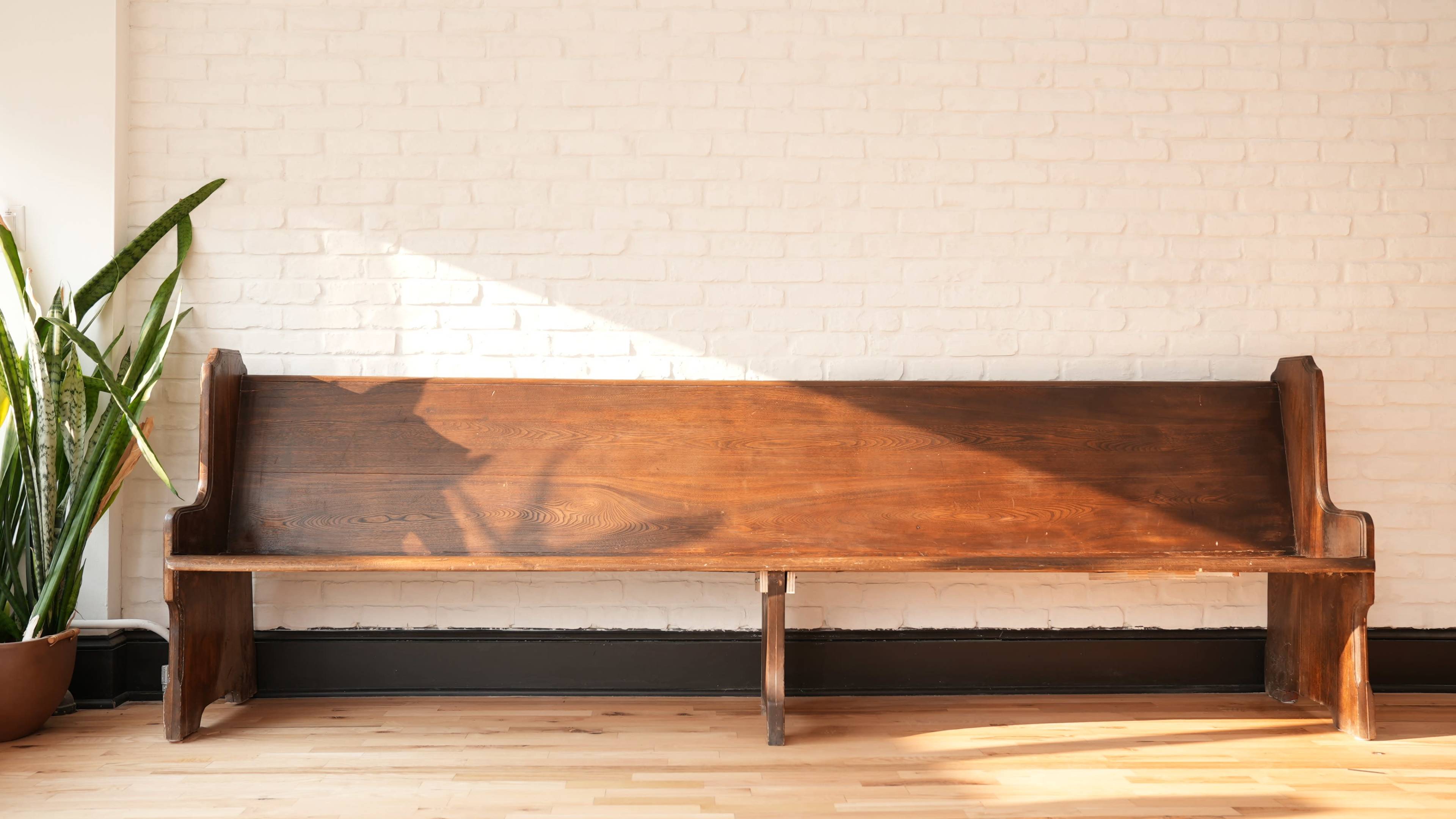 A wooden bench is positioned against a white brick wall with sunlight casting a shadow on it.