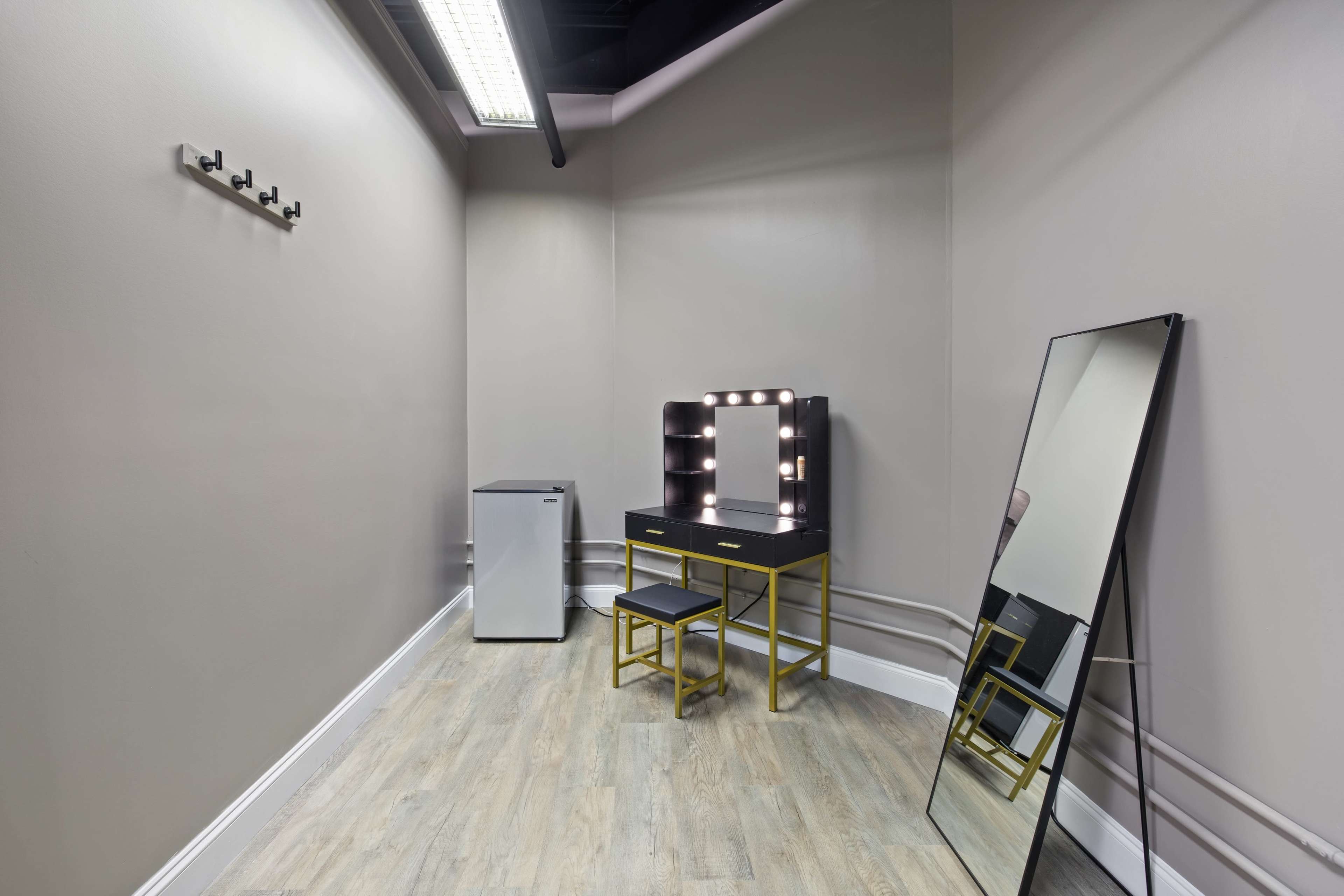 A minimalist room with a mirror, a vanity table with a lighted mirror, a chair, a small refrigerator, and wall hooks.