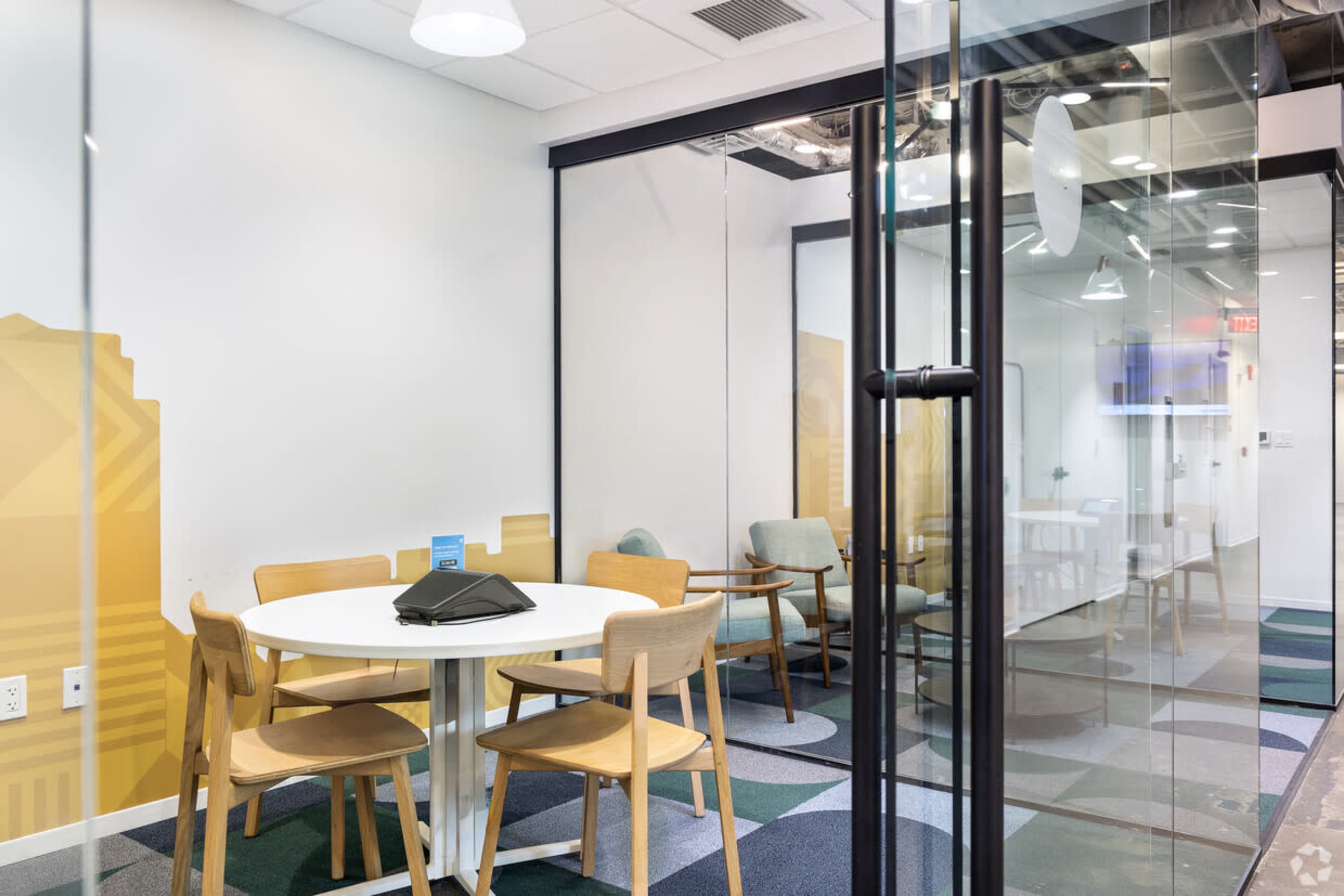A modern meeting room features a round white table surrounded by wooden chairs, with glass walls and a large television visible in the background.