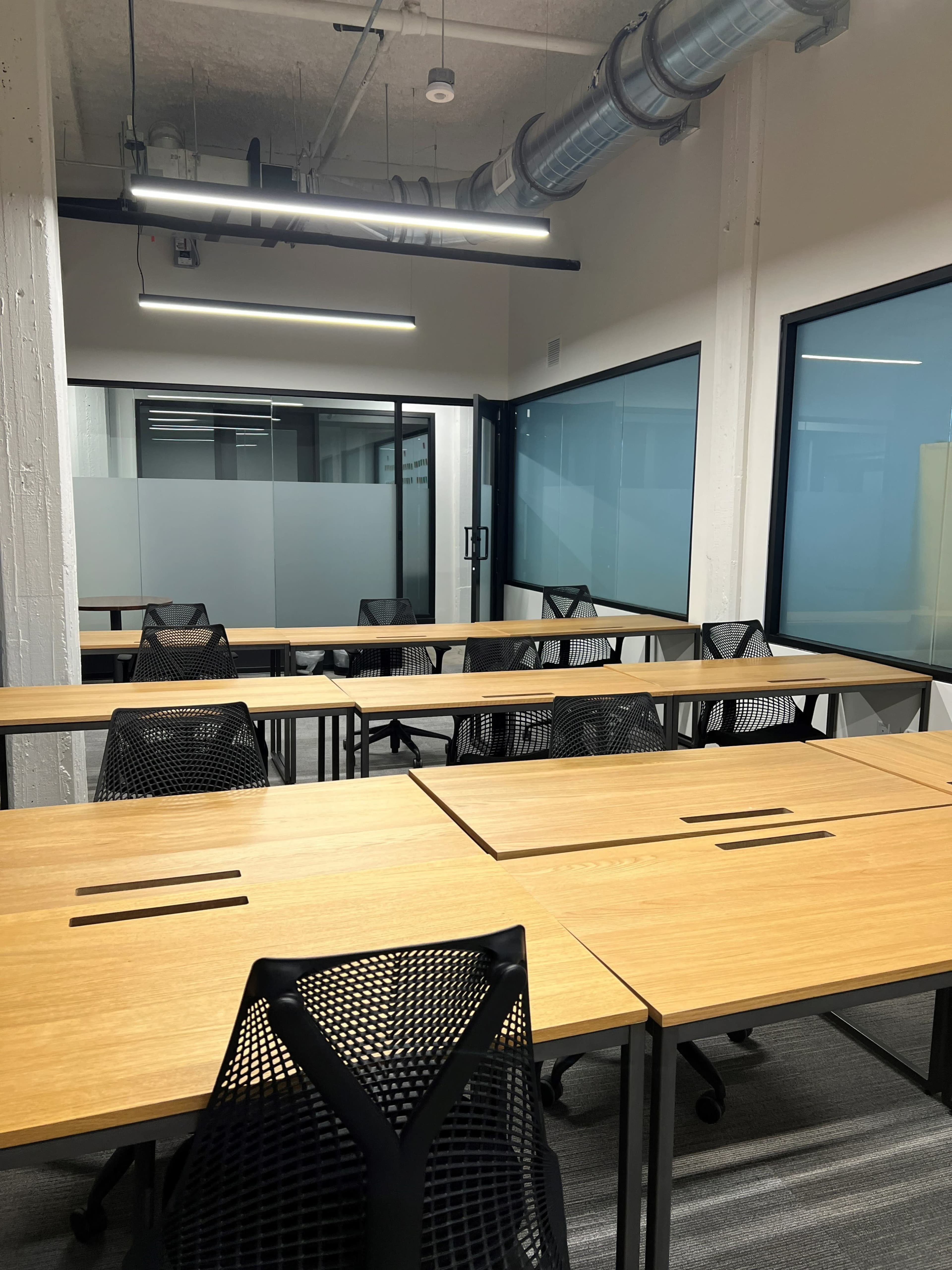 An empty modern office space with multiple wooden desks and mesh chairs arranged in a grid pattern, separated by glass partitions.
