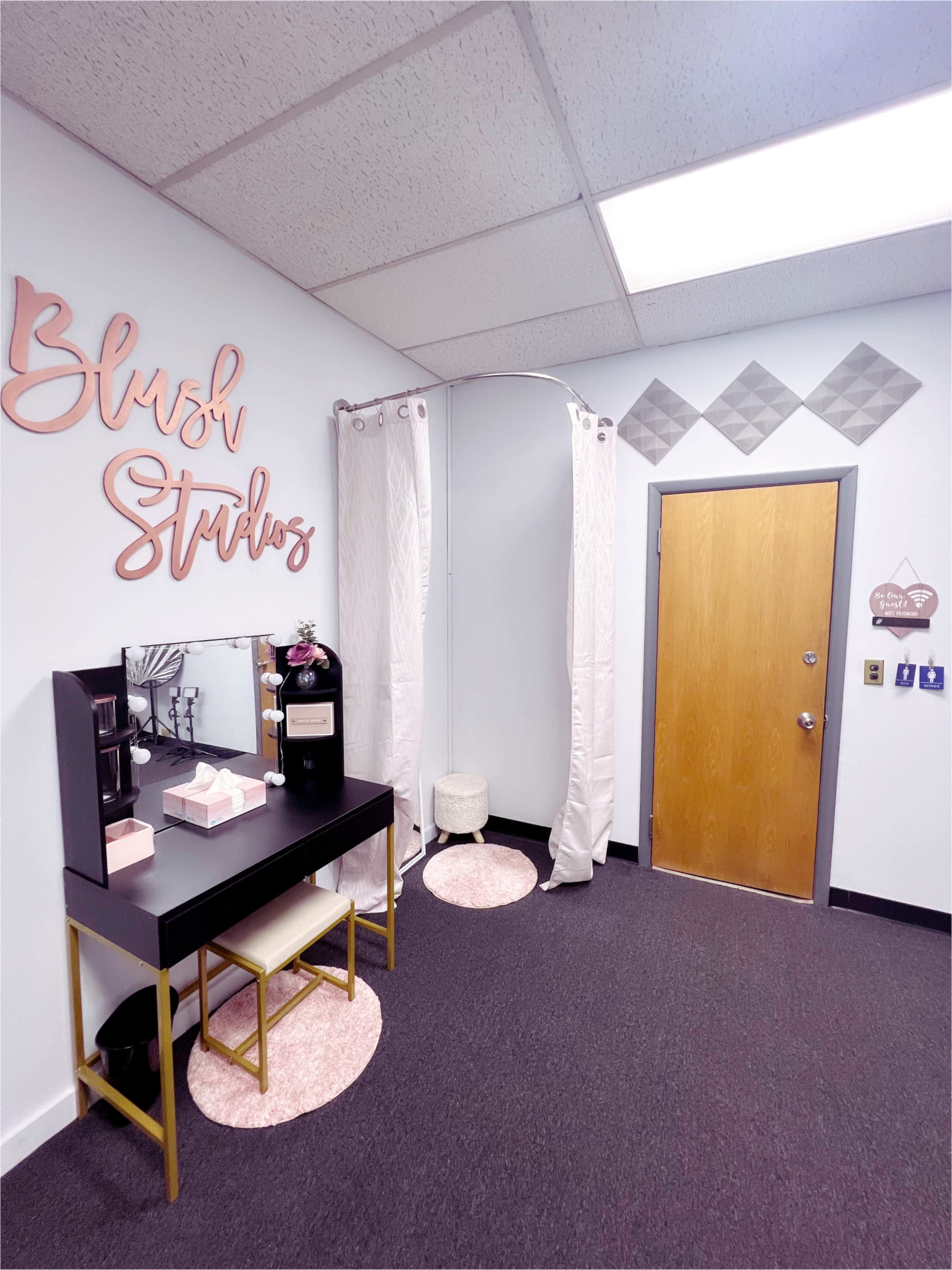 The image shows a neat beauty studio entrance featuring a black vanity table with mirror, pink accents, and a door with a decorative sign.