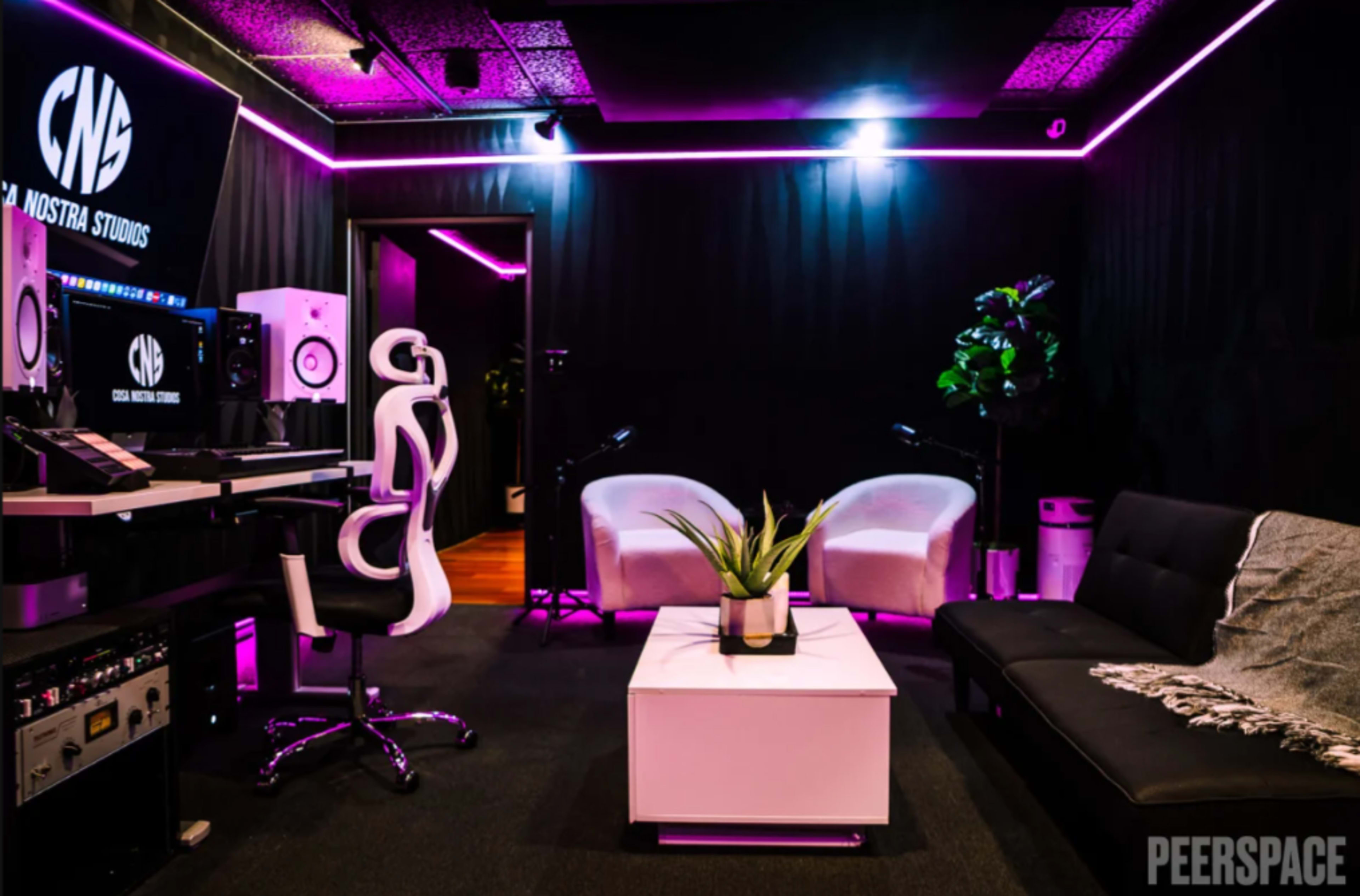 The image shows a modern recording studio with a black and pink color scheme, featuring a computer setup, two white chairs, a couch, and a small table with a plant.