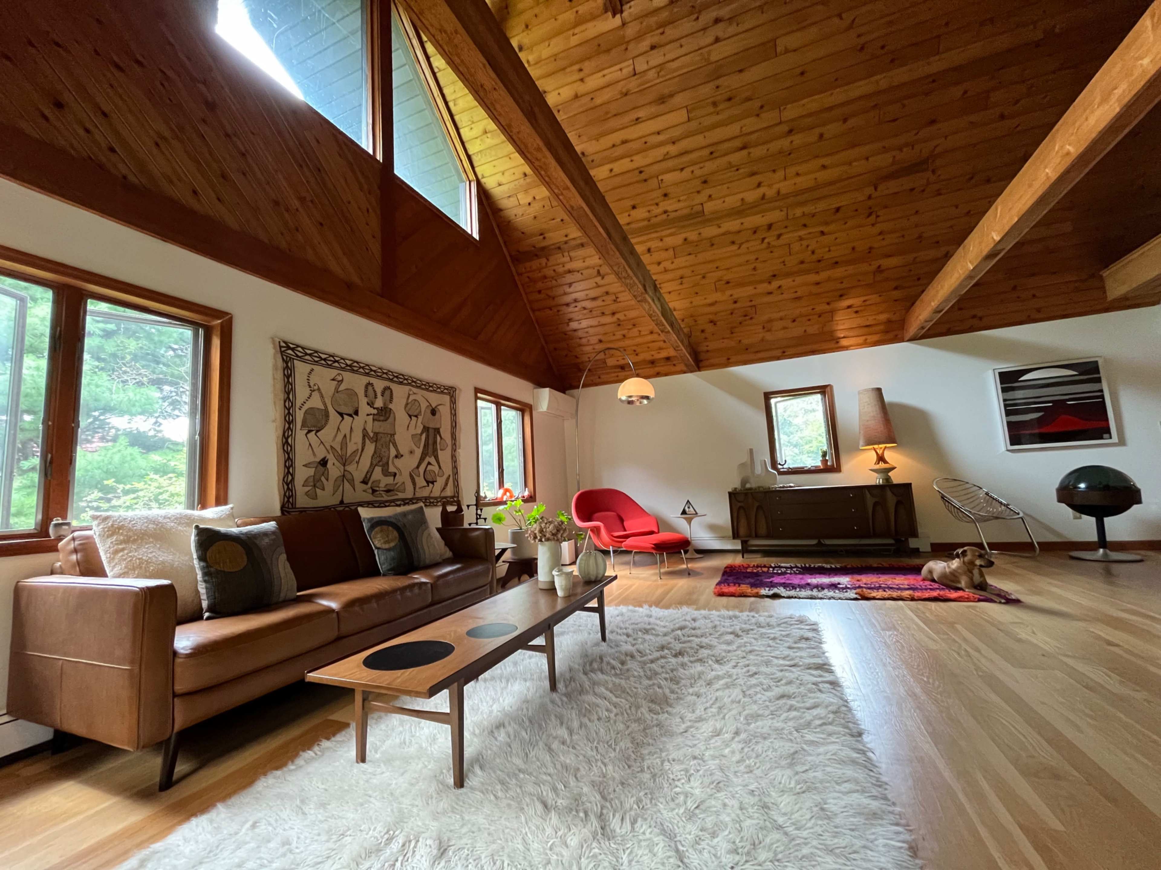 The image shows a spacious living room with wooden beams, a brown leather sofa, a coffee table, and a rug, along with large windows providing a view of the outdoors.