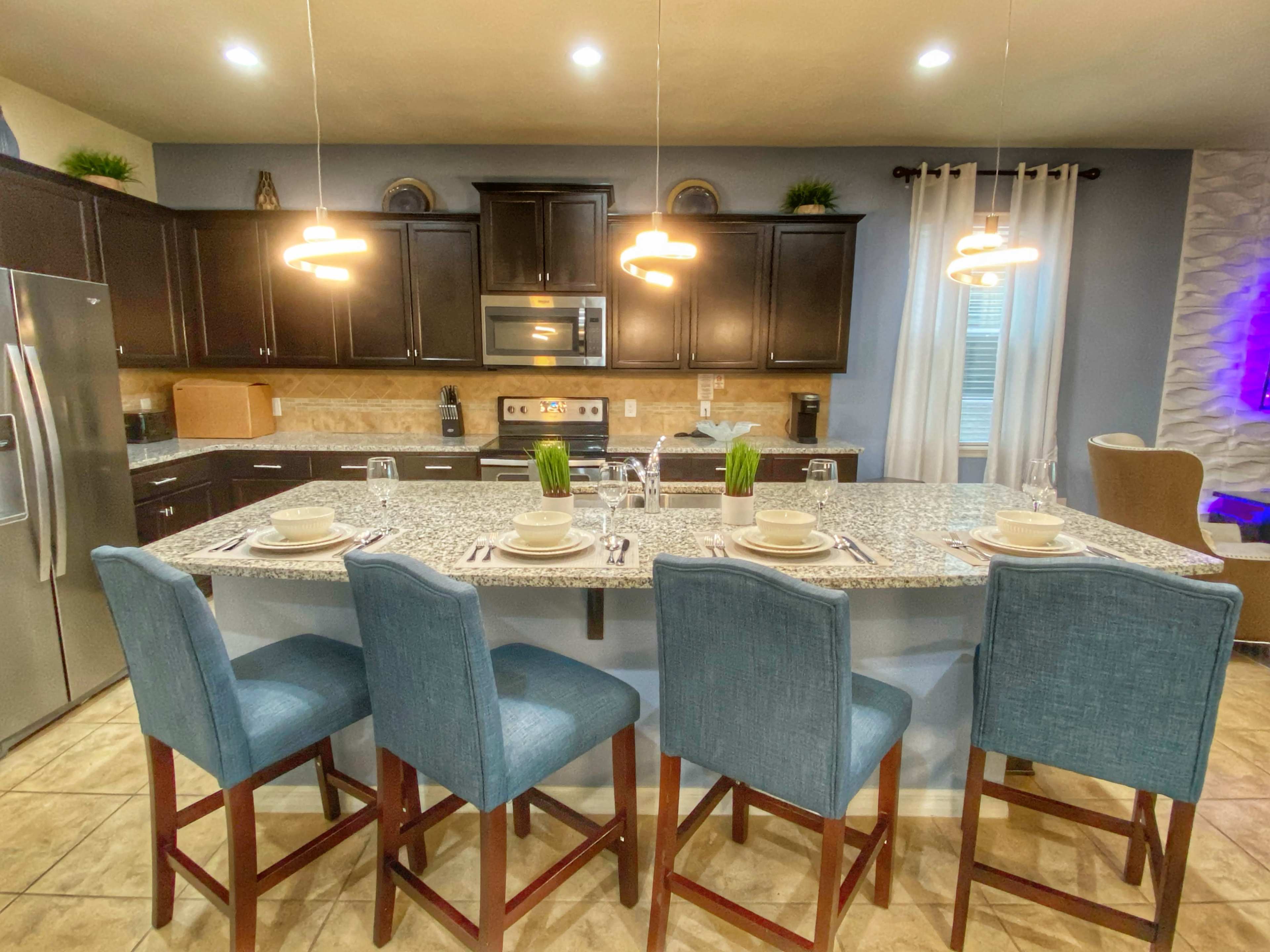 A modern kitchen features a granite countertop island with four blue bar stools, stainless steel appliances, and warm cabinetry.