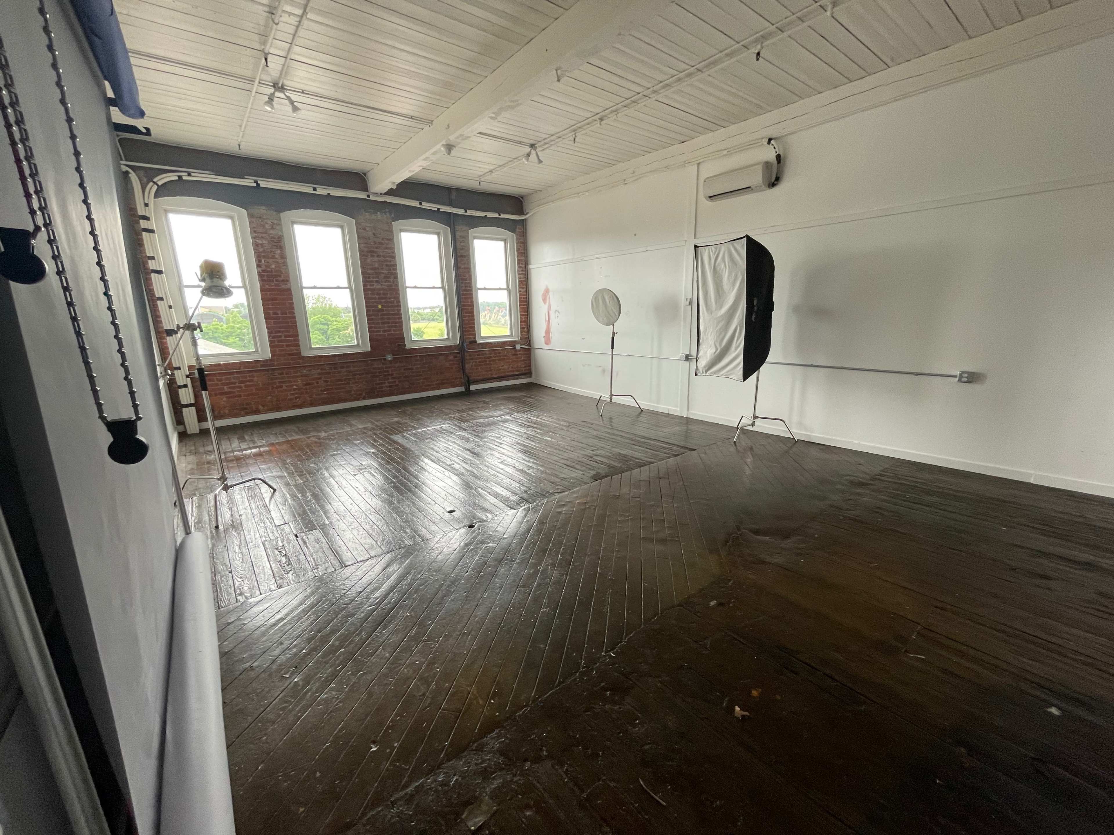 An empty photo studio with wooden floors, white walls, large windows, and lighting equipment set up in the corners.