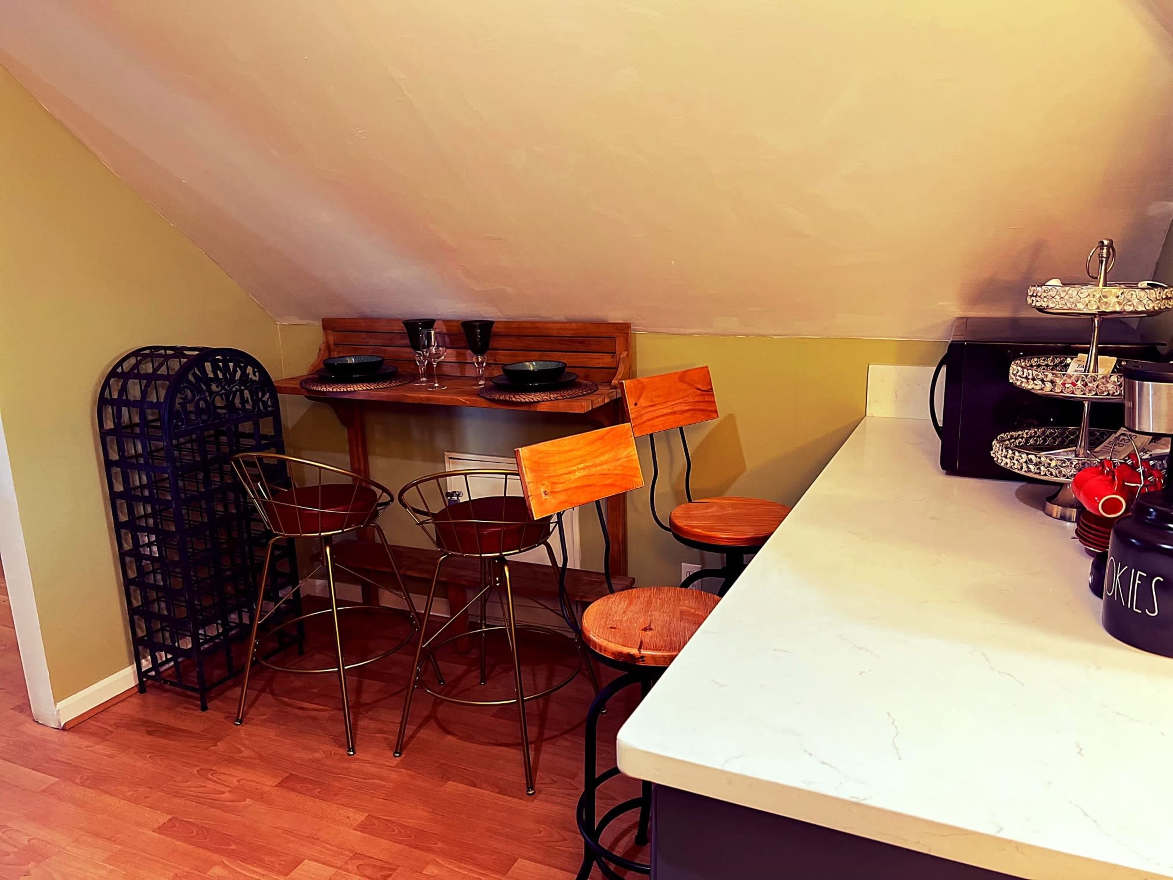 The image shows a cozy kitchen area with a counter, three wooden bar stools, and a small dining table set against a sloped wall.