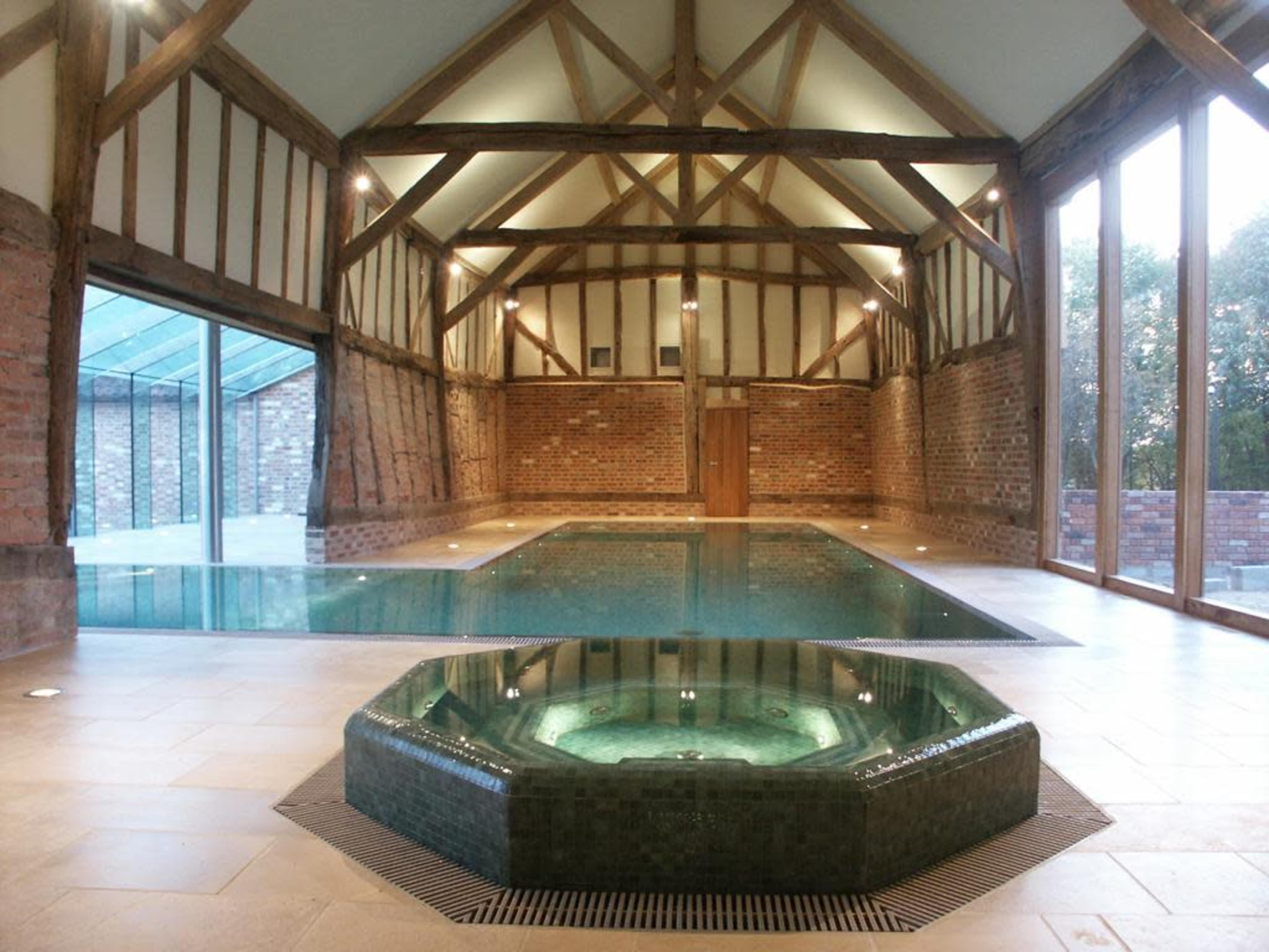 An indoor swimming pool with a sunken hot tub, set in a spacious room featuring exposed wooden beams and large glass windows.