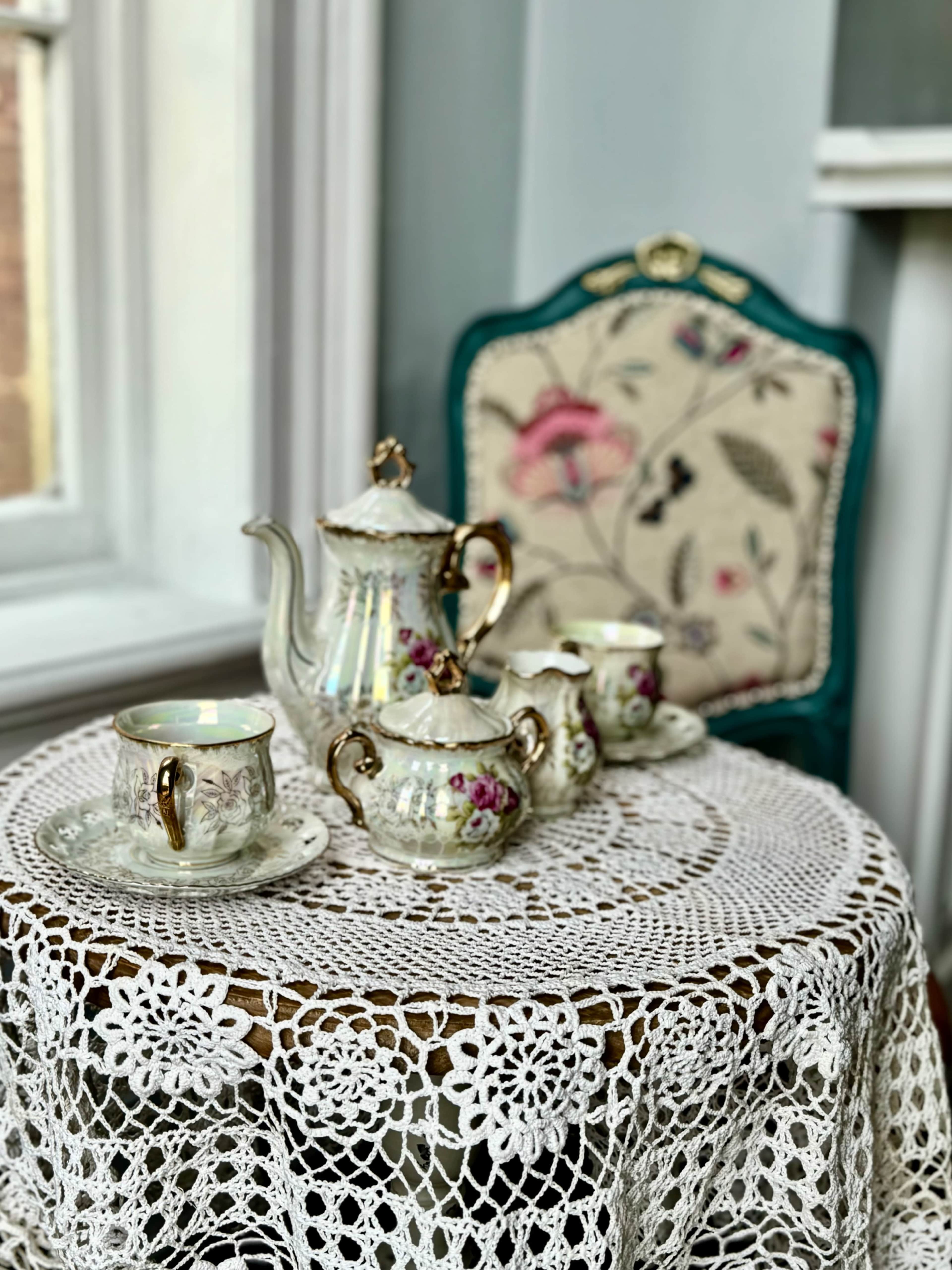 A porcelain tea set is arranged on a lace tablecloth atop a round table near a window, with a decorative chair in the background.