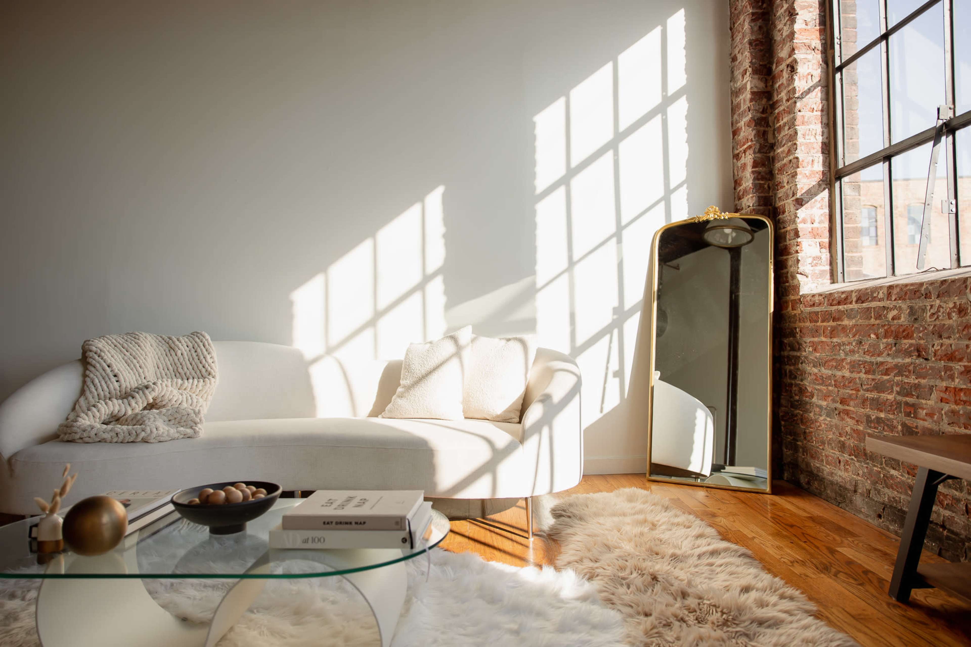 A light-filled living room features a white sofa, a glass coffee table, a decorative bowl, and a large mirror reflecting sunlight against a brick wall.