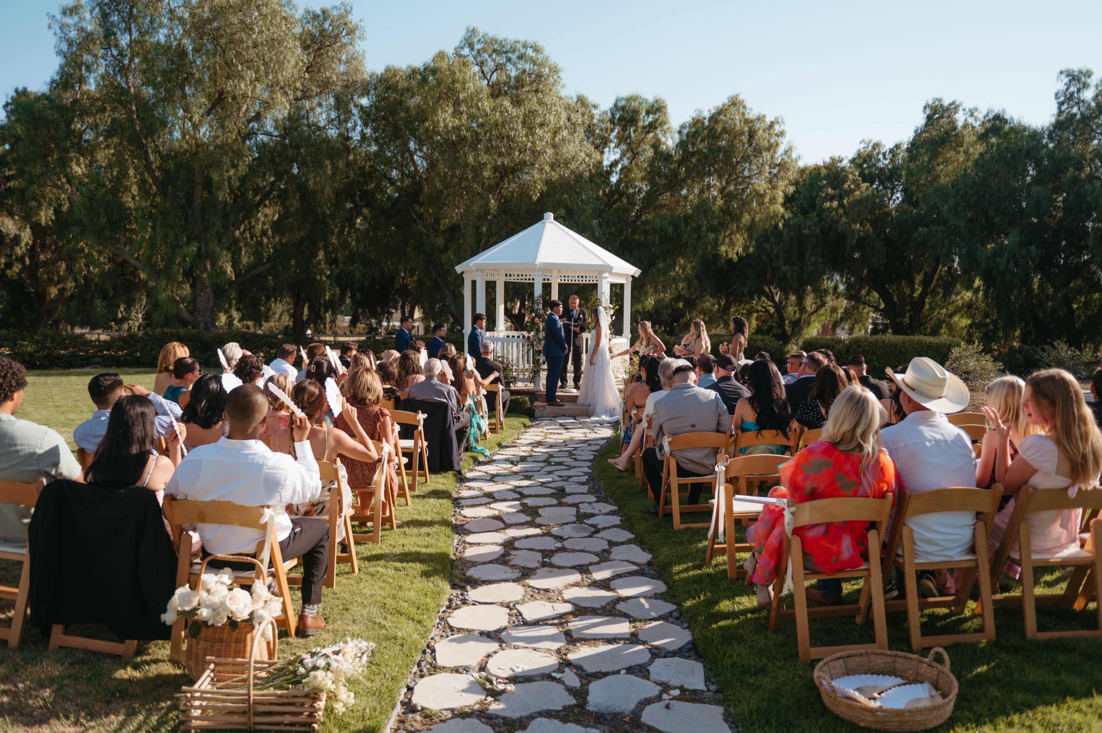 A wedding ceremony takes place outdoors in front of a gazebo, with rows of guests seated on either side of a stone path.