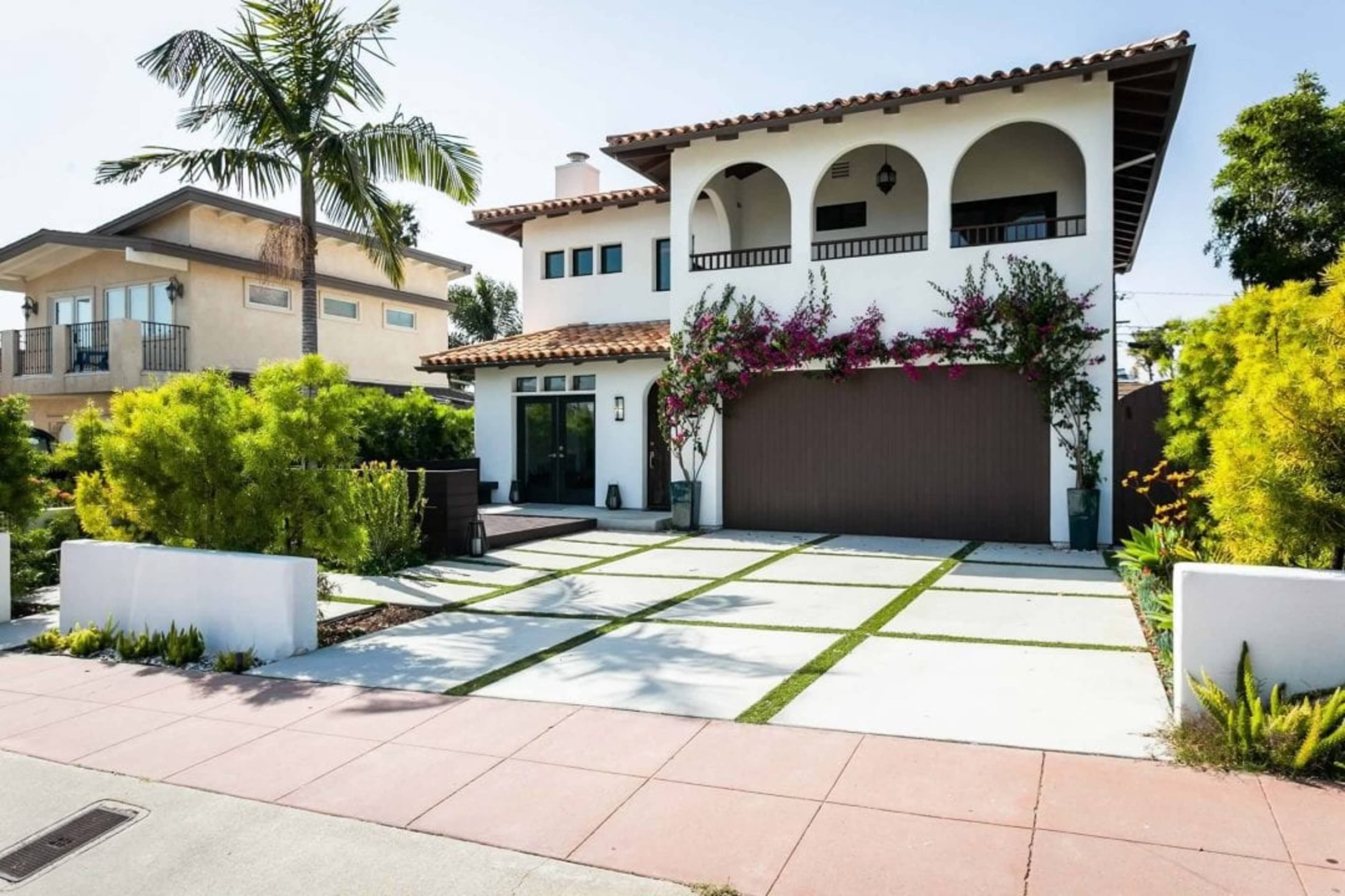 A two-story house with a tiled roof, a large garage door, and a landscaped front yard featuring palm trees and vibrant flowers.