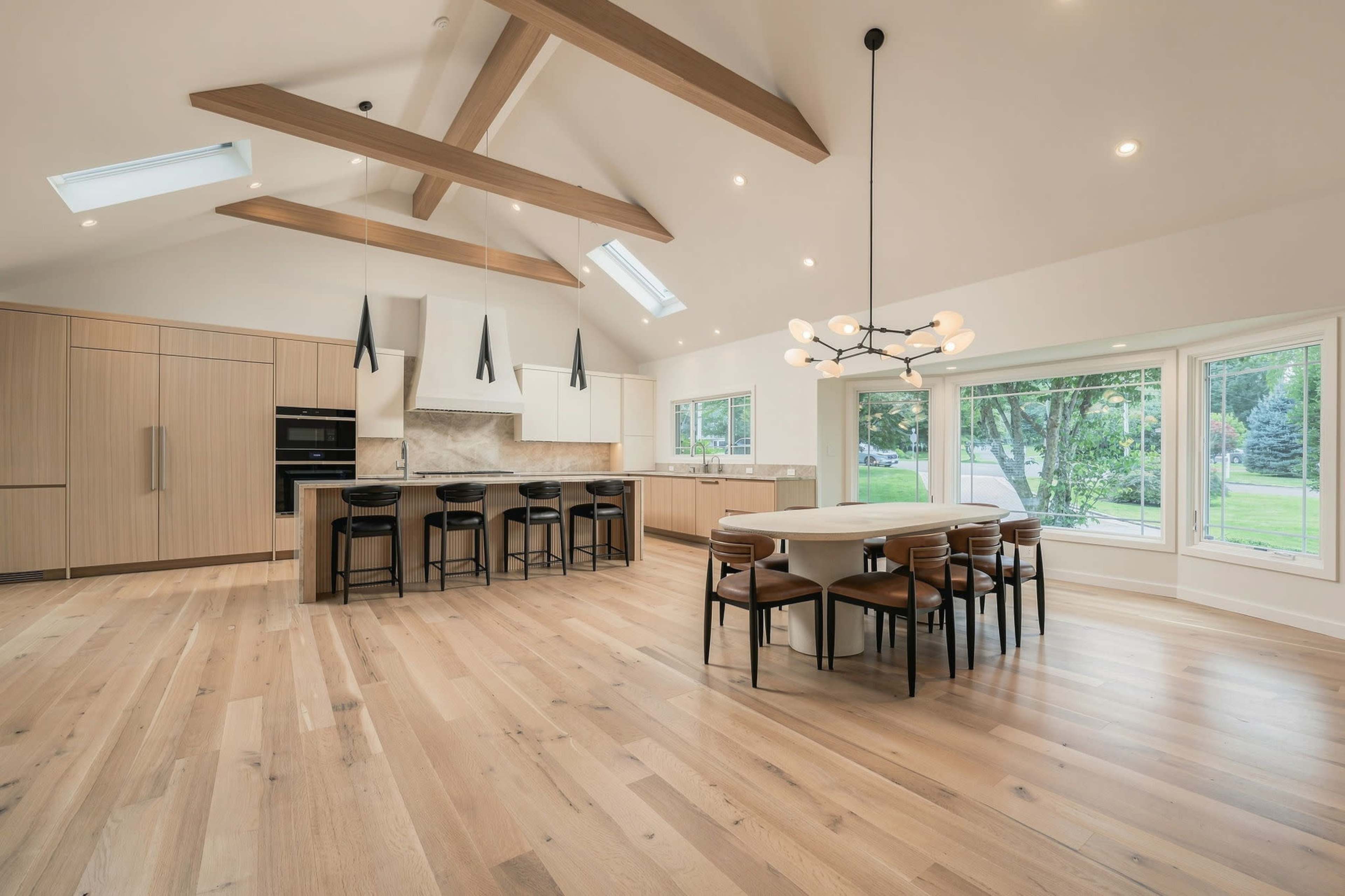The image depicts a modern kitchen and dining area featuring a spacious open layout with wooden beams, a large circular dining table surrounded by chairs, and ample natural light through large windows.