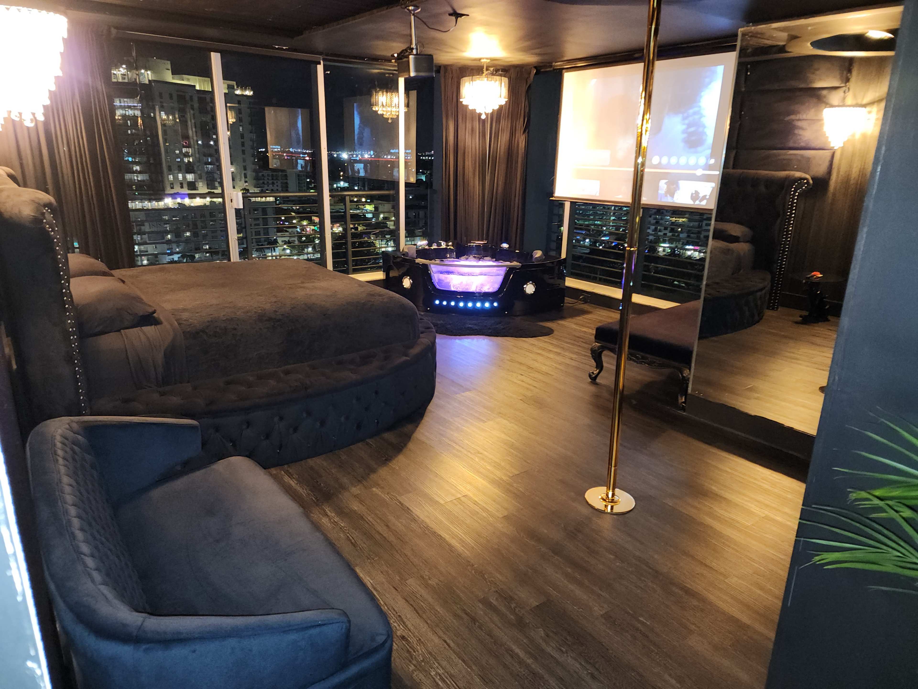 The image shows a modern hotel room with a large bed, a lounge area, a mirrored wall, and a projector screen displaying a video.