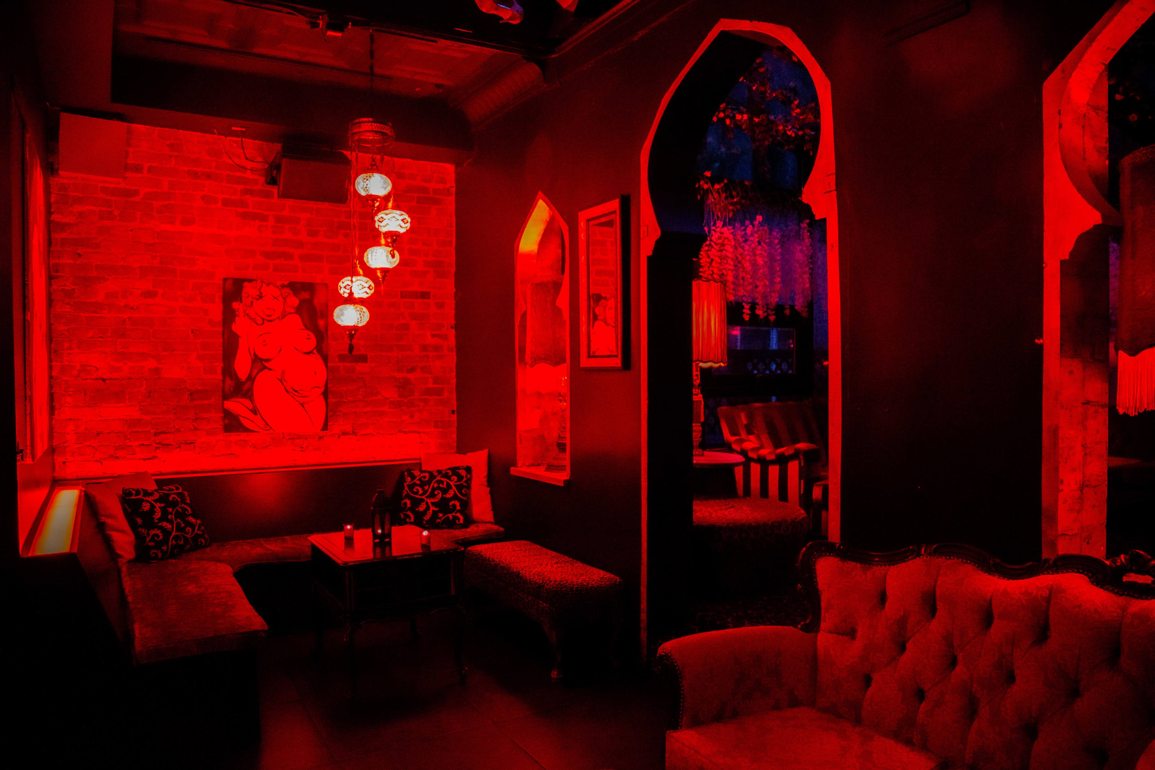 A dimly lit lounge with red lighting, featuring upholstered seating, a framed artwork on the wall, and decorative lanterns hanging from the ceiling.
