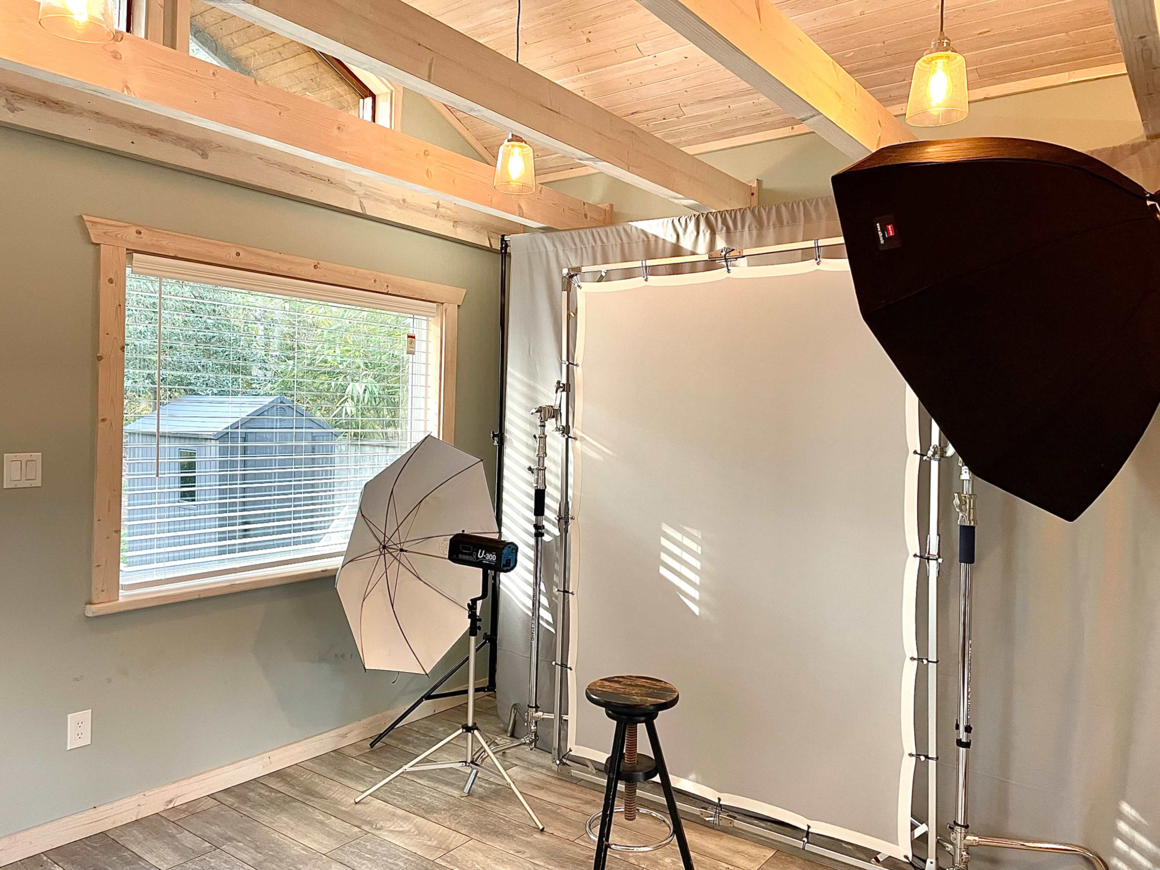 The image shows a photography studio setup featuring a large white backdrop, softbox lighting, a tripod-mounted light, and a stool positioned on a wood floor beneath wooden beams and hanging lights.