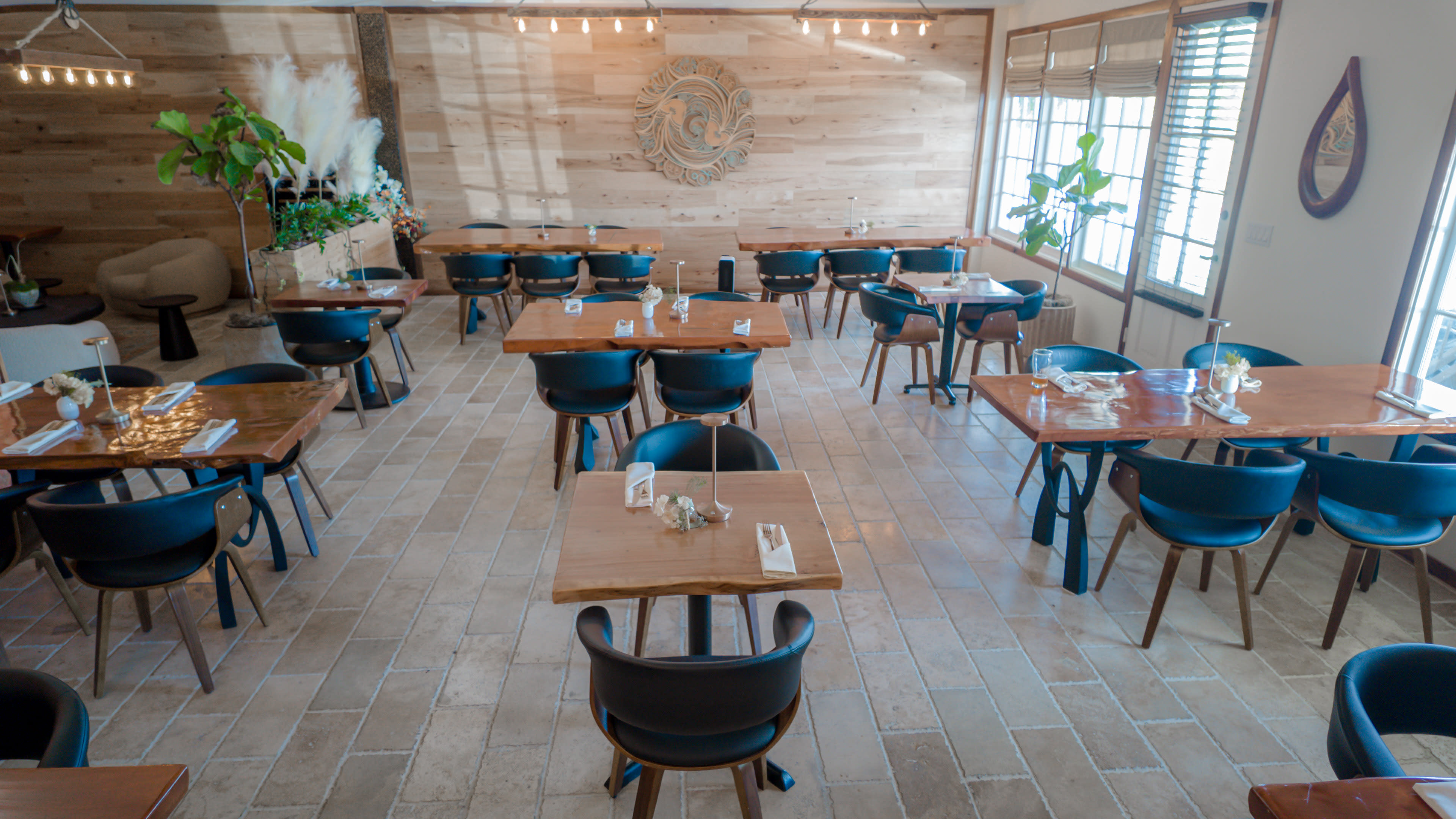 A spacious restaurant interior features wooden tables with black chairs, surrounded by light-colored walls and decorative plants.